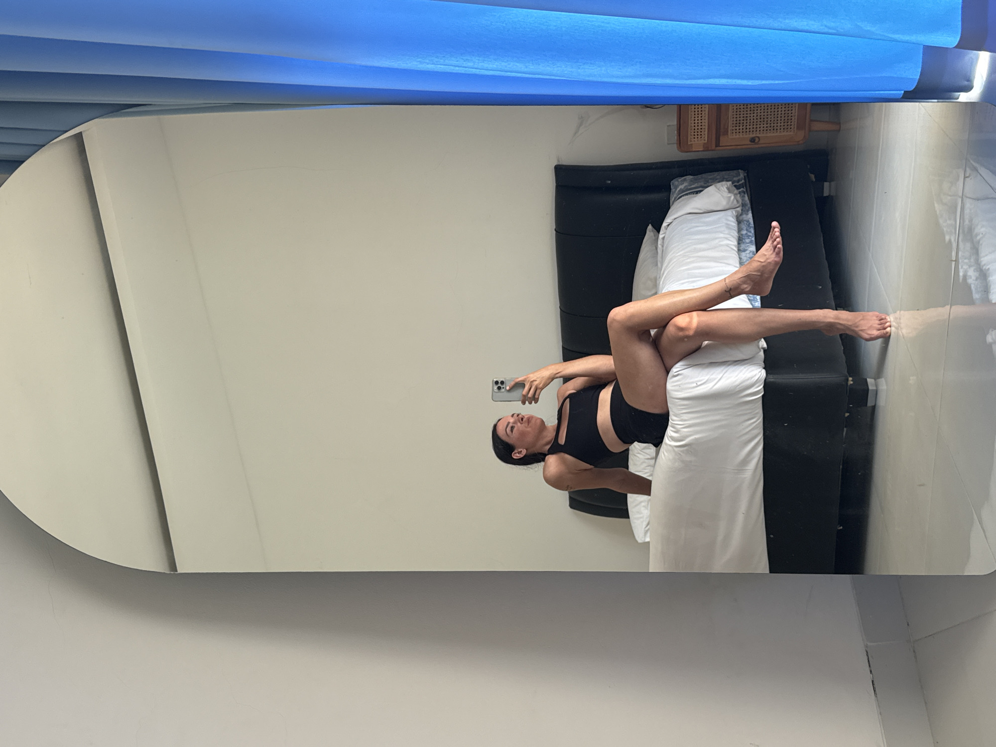 A woman in black athletic wear is captured in a mirror reflection, taking a selfie while sitting on the edge of a bed in a room in Mengwi, Indonesia. She is positioned on the right side of the bed, her legs crossed and extended. The room features a dark headboard, white bedding, and a blue curtain on the right side. A small wooden nightstand is visible next to the bed. The floor is tiled and reflects the scene.