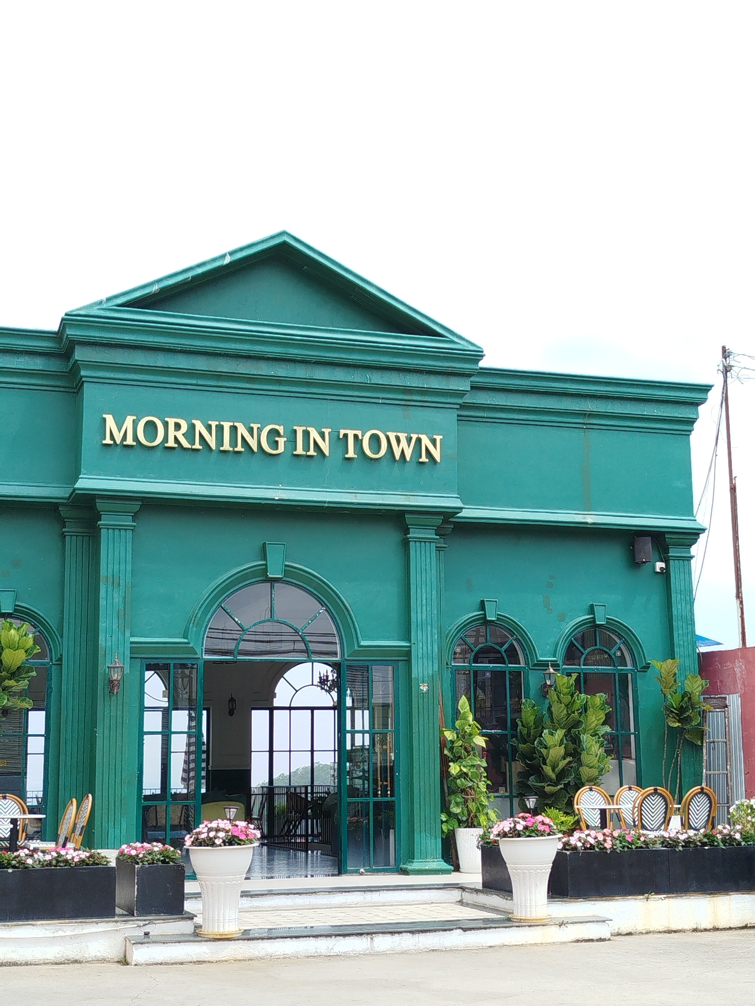 The image displays the exterior of a building named "MORNING IN TOWN," likely a cafe or restaurant, located in Da Lat, Vietnam. The building is painted a distinctive teal-green and features classical architectural elements, including a prominent pediment at the top, decorative columns framing the main entrance, and detailed cornices. Gold-colored letters spelling "MORNING IN TOWN" are affixed to the facade above the entrance.
The front of the establishment boasts large arched windows and a central arched glass doorway, allowing views into the interior. An inviting outdoor seating area is visible, furnished with bistro-style chairs featuring woven backs—some in a black and white pattern, others in a lighter tone—arranged around small tables. White fluted planters and black rectangular planters overflowing with pink flowers line the steps and the outdoor seating area, adding a touch of color. Large potted green plants are also placed strategically next to the windows.
The scene is brightly lit by diffuse, possibly overcast, daylight, suggesting a morning or early afternoon setting, aligning with the establishment's name. No people are present, indicating a quiet moment. A black security camera is mounted on the upper right side of the building, and a utility pole with wires is visible further right, hinting at the surrounding environment. The overall impression is one of charming tranquility and a welcoming atmosphere.