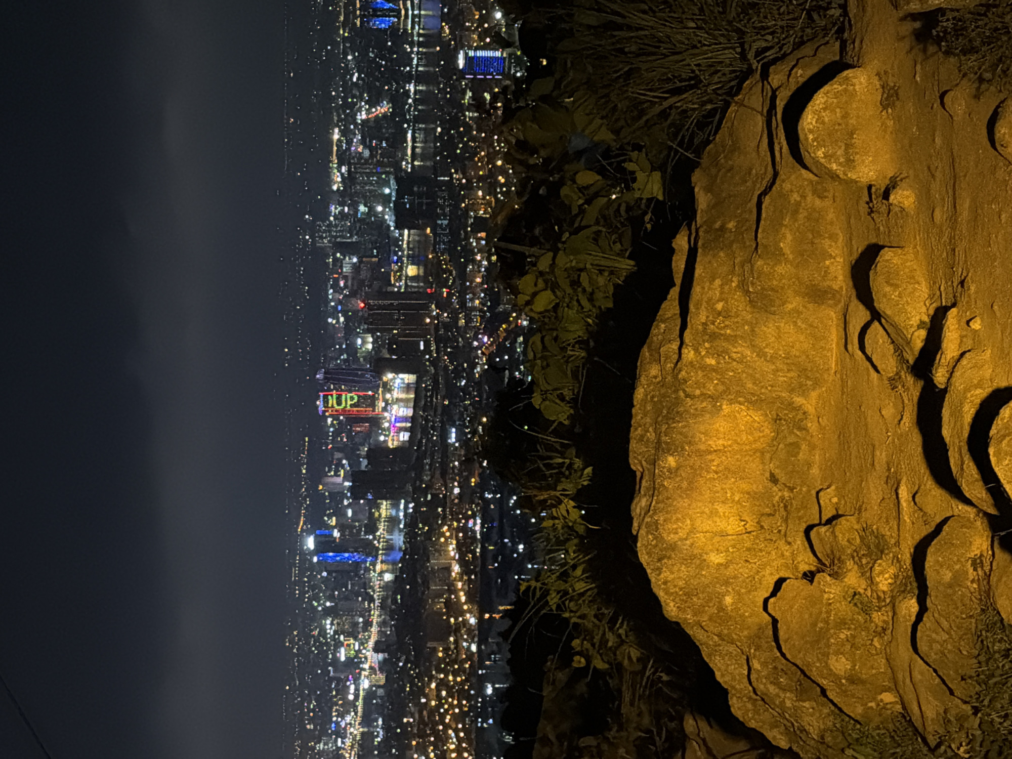 This nighttime image captures a cityscape of Đà Nẵng, Vietnam, viewed from an elevated position, possibly a mountain or high vantage point. The city lights illuminate the scene, with buildings and streets visible in detail. In the foreground, there's a large rock formation, adding depth to the composition. The sky is dark, suggesting a clear night. The photograph seems to be taken from a stationary position. There are no people visible. The word “DUP” is visible atop a lit building in the city below.