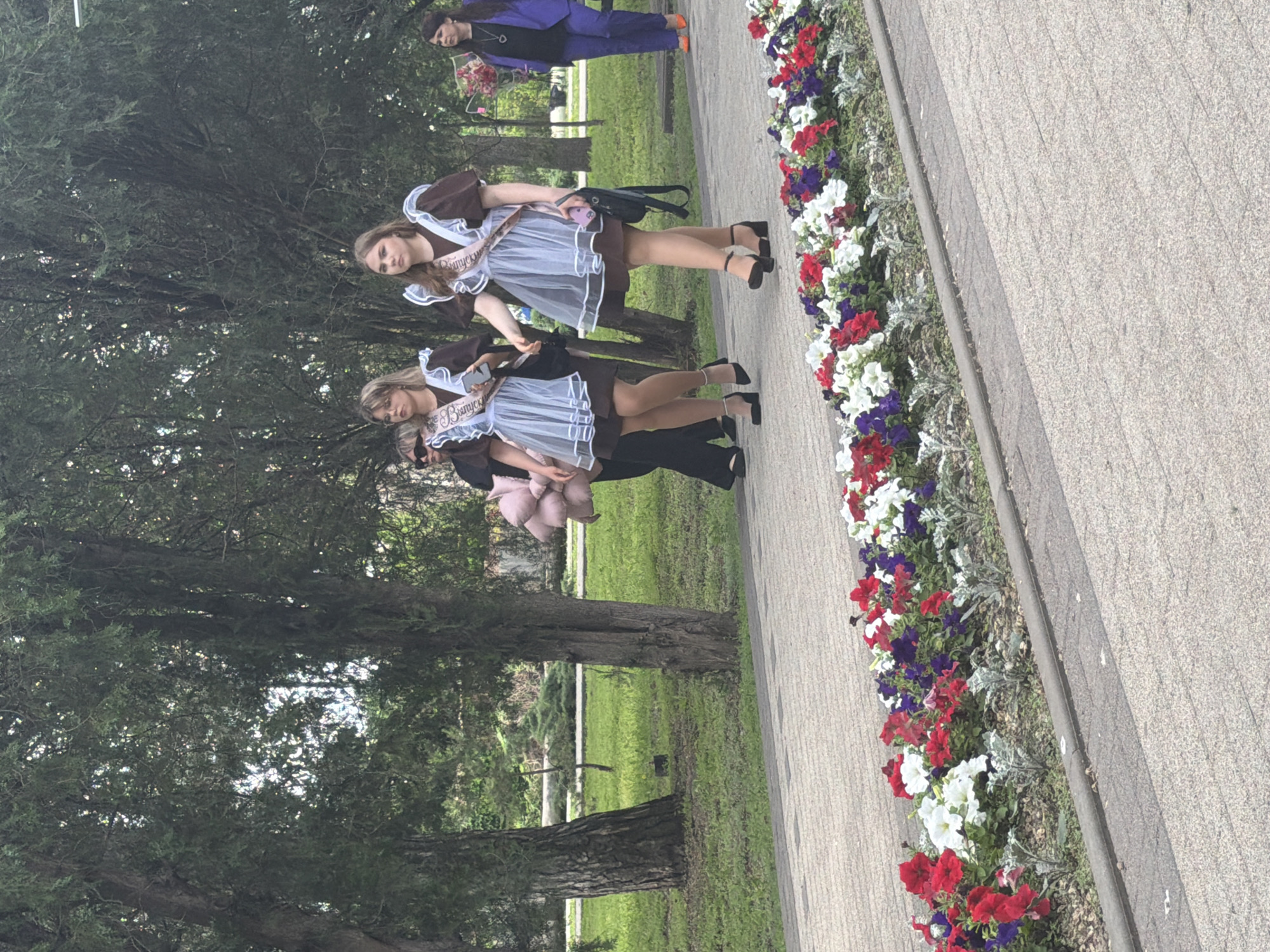 The image shows a paved walkway in Leningradskaya, Russia. Along the walkway is a flowerbed with red, white, and purple flowers. Two young women are walking side-by-side. They wear traditional school dresses and " выпуск " sashes. A person in a blue suit with a bouquet of flowers walks ahead of them. A tree line borders the walkway and a grassy area stretches behind it.