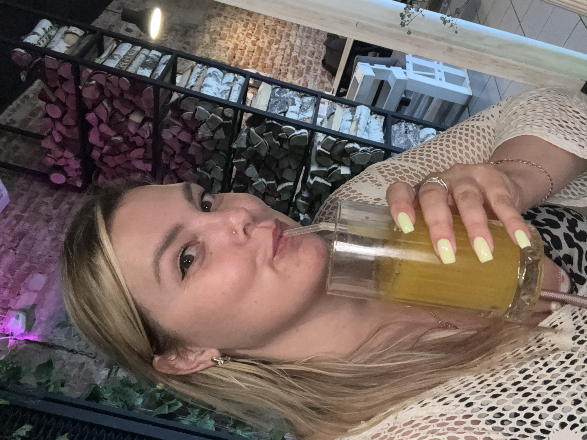 A blonde woman with light skin and makeup is holding a glass of yellow liquid with a straw and drinking from it. She is wearing a white, open-knit top and has light yellow nail polish. She is also wearing a ring on her right hand and a bracelet. The background shows a brick wall, shelves stacked with logs, and some greenery. The lighting suggests an indoor setting, possibly a restaurant or bar. The image does not provide any information or visual cues that would allow for identification of the specific location in Chaikovsky, Russia.