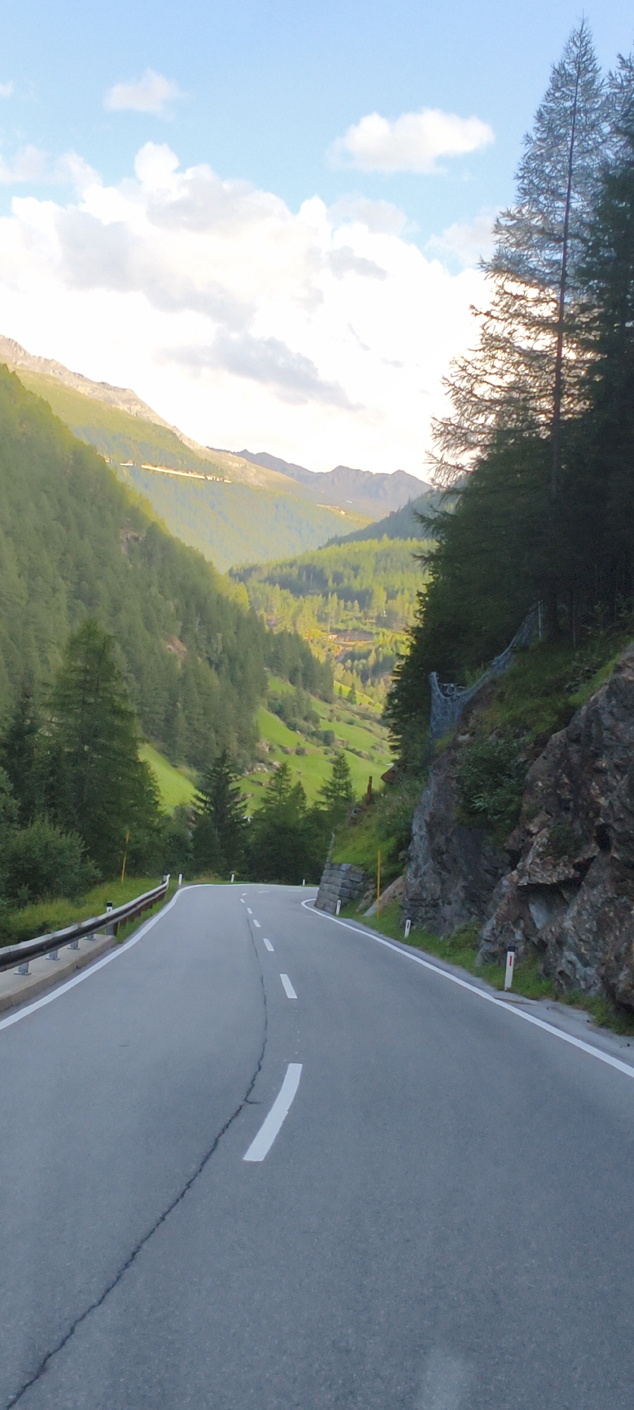 The image depicts a winding asphalt road leading into a lush green mountain valley in the Austrian Alps, likely near Sölden. The road is marked with white dashed lines and has a guardrail on the left side. The valley walls are densely covered with evergreen trees, and in the distance, rolling hills and rugged mountain peaks are visible under a bright blue sky with scattered white clouds. The scene conveys a sense of natural beauty and solitude.