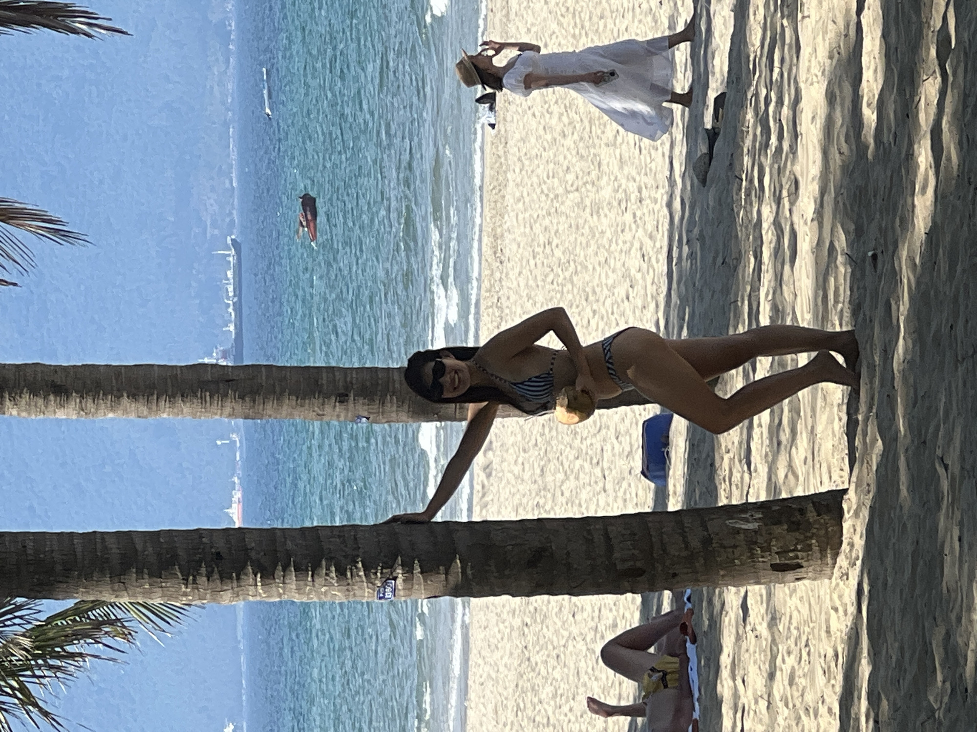 The image depicts a sunny day at My Khe Beach in Da Nang, Vietnam. In the foreground, a woman in a striped bikini stands beside a palm tree, holding a coconut. She is wearing sunglasses and smiling at the camera. Behind her, another woman in a white dress and a straw hat walks along the beach. To the left, partially visible, is a person lying on a towel. The turquoise ocean stretches to the horizon, dotted with a few boats and ships. Palm trees line the sandy beach, casting shadows on the ground. The overall atmosphere is one of relaxation and enjoyment of a tropical beach setting.