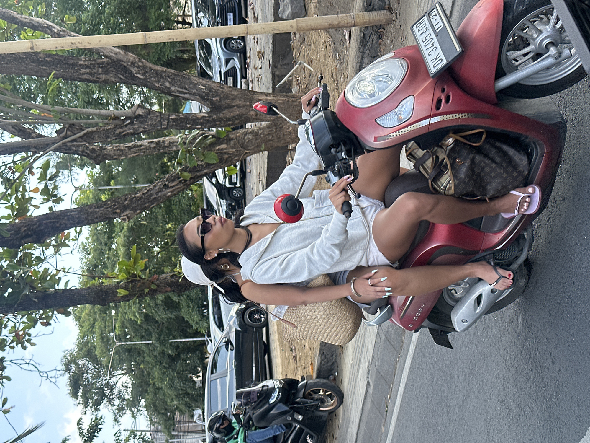Two young women ride on a red scooter on a street in North Kuta, Indonesia. The woman in front is driving, wearing a white hoodie, white shorts, sunglasses, and a white baseball cap. She has long, dark hair. The woman behind her wears a white tank top and denim shorts. She has long, dark hair and is wearing a straw bag. The scooter has a license plate that reads "DK 3405 ADR 11-27". In the background, several cars are parked and trees line the street. The sky is partly cloudy.