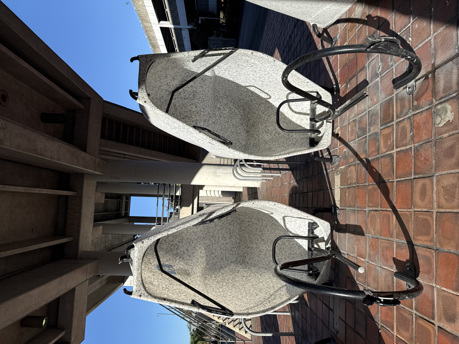 Two futuristic, white, speckled bike racks sit on a red brick walkway. Their black metal frames cast long shadows. Above, concrete architecture with visible stairs and pipes creates a layered scene, punctuated by a patch of bright blue sky. A traditional metal bike rack stands behind the unique ones.