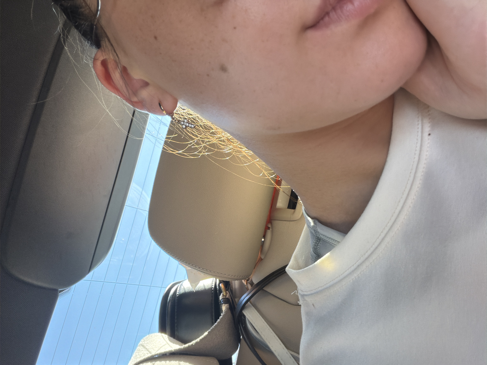 A close-up, over-the-shoulder shot captures a person's ear and cheek, illuminated by warm sunlight. The individual appears to be inside a vehicle, with a tan leather car seat and the back window visible. The car is parked in Simila, Romania, on a bright, clear day. A white t-shirt is worn by the subject, and delicate gold earrings adorn their ear. The overall scene is serene and relaxed.