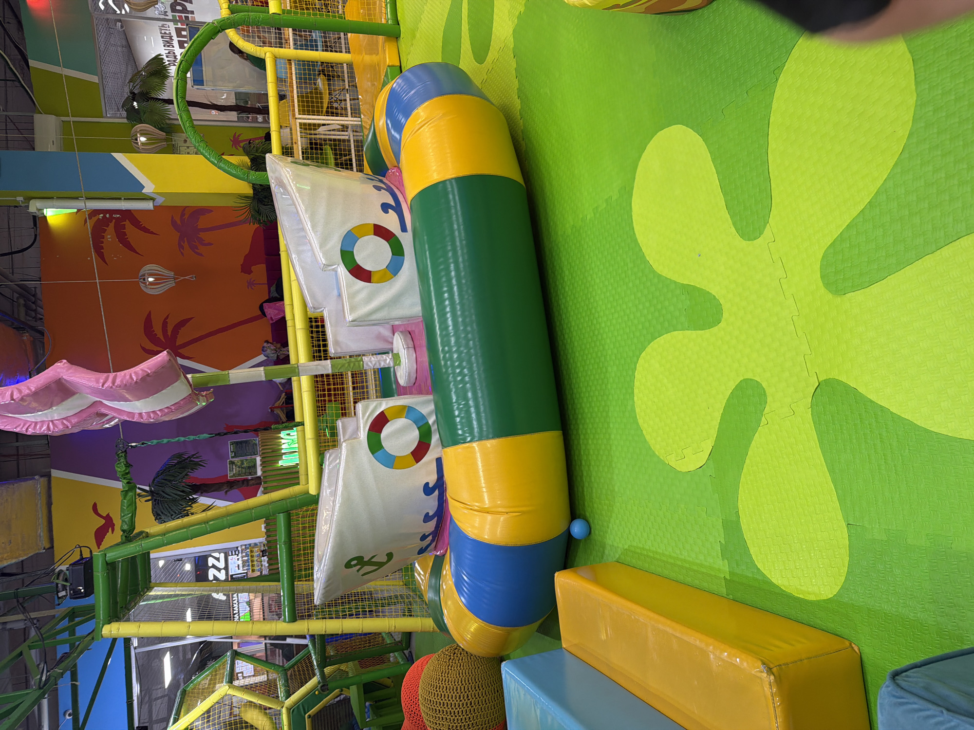 A pirate ship-themed inflatable structure is central to the image, positioned on a green, puzzle-piece mat floor in what appears to be an indoor children's play area. The inflatable ship is multi-colored, featuring sections of green, yellow, and blue, with a white hull adorned with waves and a life preserver graphic. A pink and white striped sail rises from the ship. Surrounding the inflatable are various soft play blocks in yellow and blue. In the background, a brightly colored mural depicts a tropical scene with palm trees and a bird. Sections of a climbing frame and yellow safety netting are also visible. A small blue ball rests on the floor near the inflatable. The scene is well-lit, typical of an indoor entertainment venue.