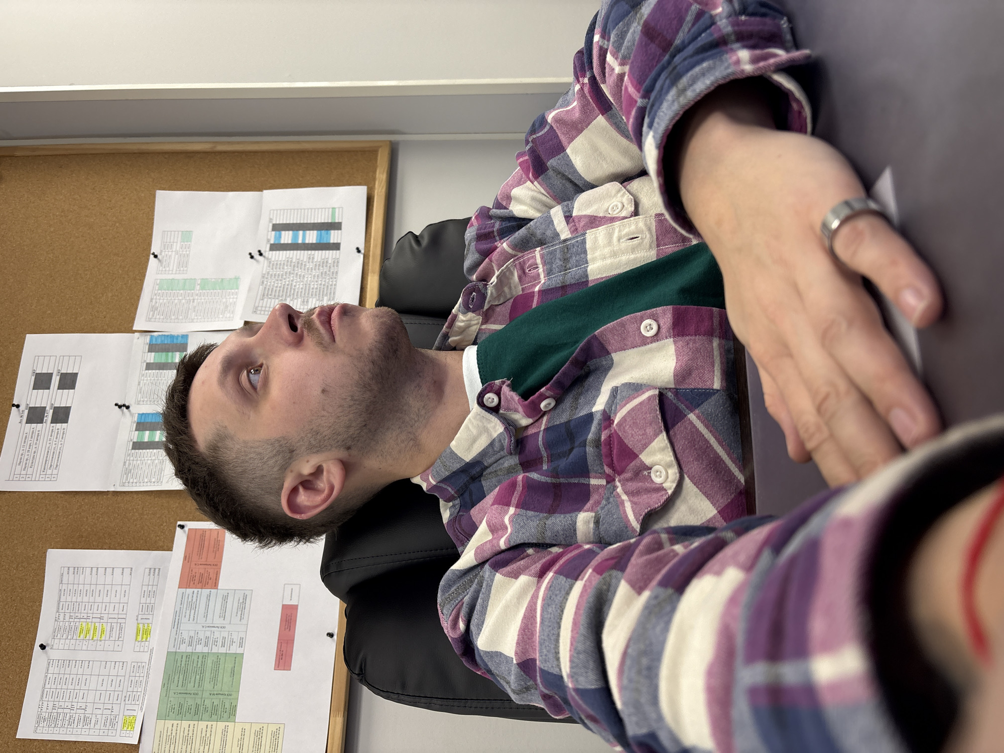 A man with short, dark hair is sitting in a black office chair. He is wearing a purple and white plaid shirt over a green shirt. He is looking up and to his left. His right hand is resting on a gray surface. He is wearing a ring on his right ring finger. A corkboard with several sheets of paper pinned to it is visible in the background. The documents appear to be printed tables with colored highlights. The location is not discernible from the image and Saint Petersburg, Russia, cannot be confirmed.