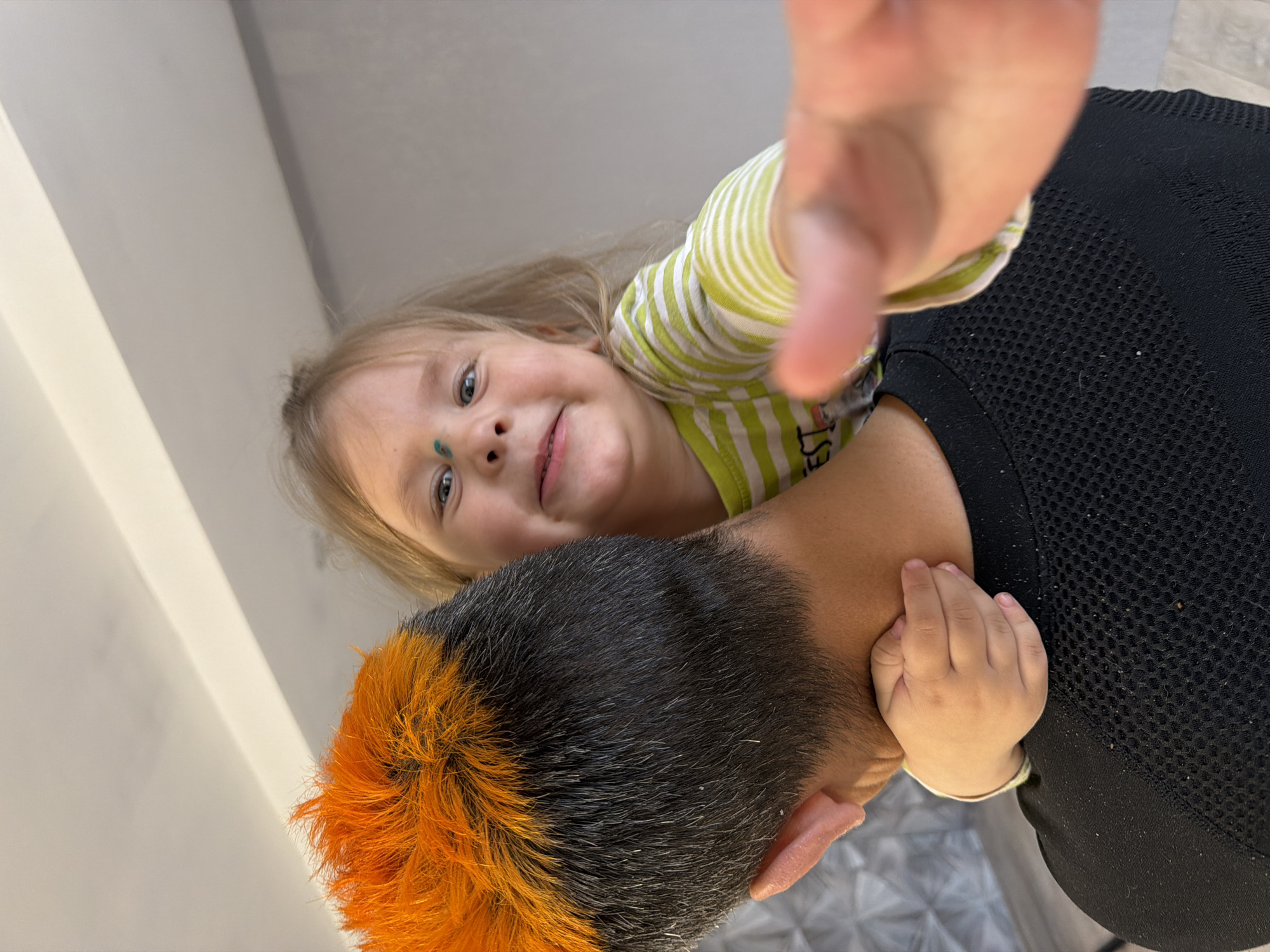 A child with blonde hair is being held up by an adult with black and orange dyed hair. The child has a green mark on their face and is wearing a green and white striped shirt. The adult is wearing a black mesh top. A hand is reaching out towards the camera. The location may be Chaikovsky, Russia.