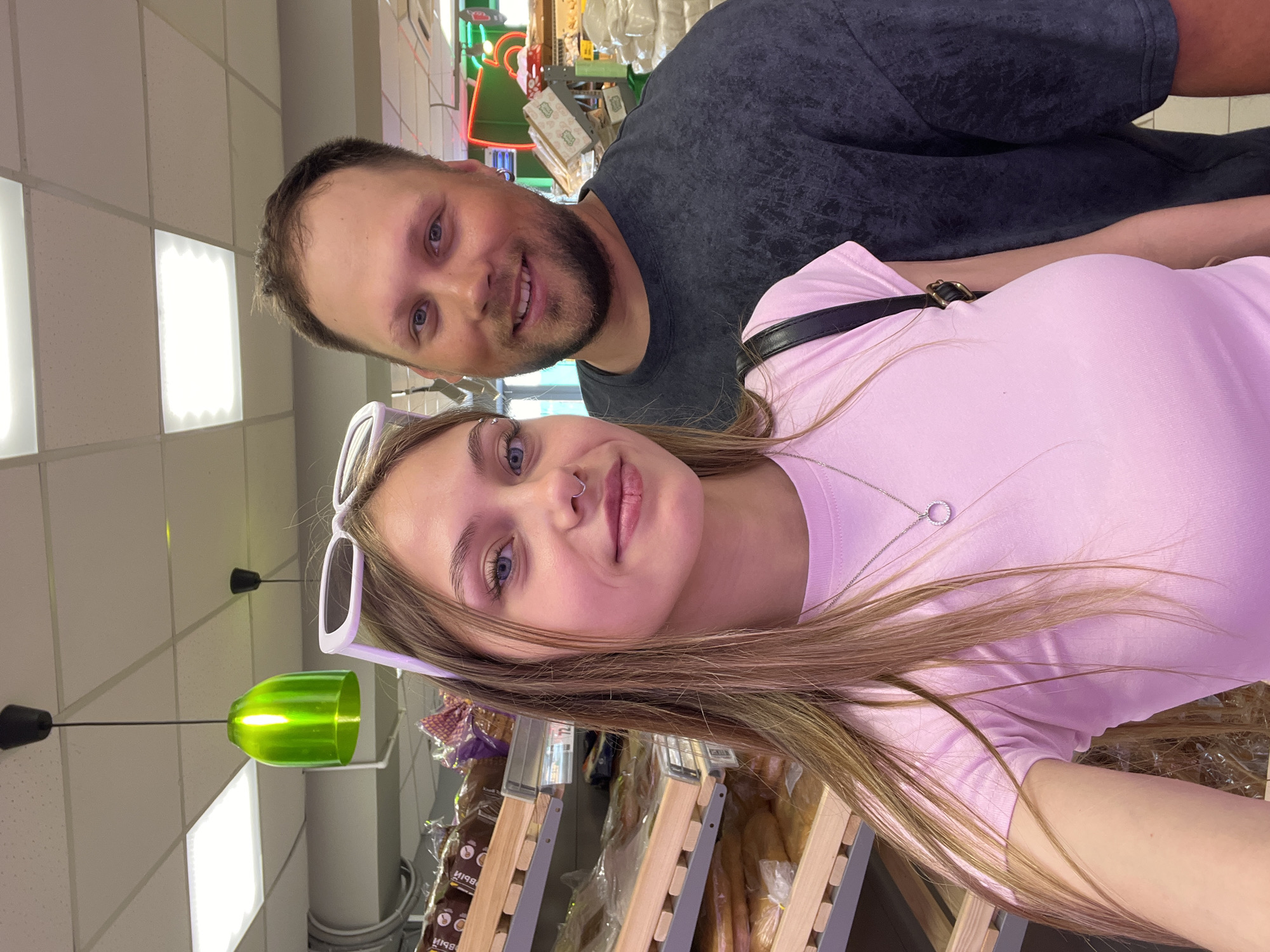 A selfie features a man and a woman in what appears to be a store setting. The woman has long, light brown hair, blue eyes, and wears white sunglasses atop her head. She has a nose piercing and a pink top with a silver necklace. The man, positioned behind her, has short brown hair and a beard, wearing a dark grey shirt.
The background reveals a retail environment. There's a display of what appears to be bread in racks. The ceiling is visible with lighting fixtures. Considering the lighting and merchandise, the setting seems to be a bakery or a grocery store. Without additional information, it's impossible to confirm the location in Фоки, Russia.