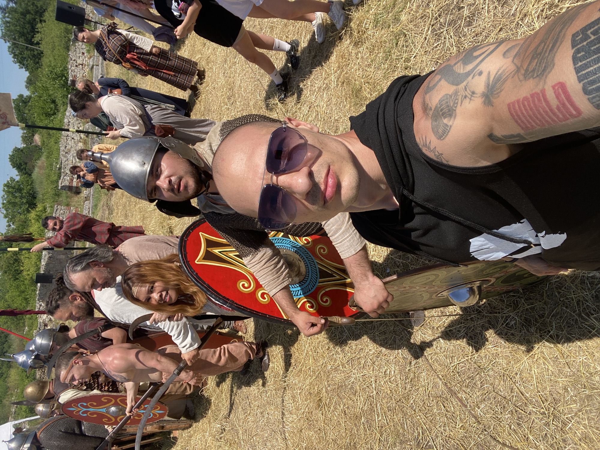The image captures a historical reenactment event in Gigen, Bulgaria, under direct sunlight on dry, straw-covered ground. Ancient stone ruins and sparse green vegetation are visible in the background.
In the immediate foreground, a bald man with purple-tinted aviator sunglasses is taking a selfie, looking directly at the camera. He wears a black sleeveless hoodie, revealing extensive tattoos on his left arm, including red geometric lines, a microphone, and tropical flora. His right hand holds the handle of a circular shield, dark green with gold and black patterns, positioned vertically.
Behind the selfie-taker, partially visible, is a man wearing a silver Roman-style helmet and chainmail, with a beard, looking towards the camera. To his left, a line of male reenactors stands. These individuals are dressed in historical military attire, including metal helmets, chainmail, and tunics. They carry various weapons such as swords and polearms, and hold rectangular or oval shields decorated with distinct patterns (e.g., red with blue/silver designs, or red with gold/brown motifs). One bare-chested male reenactor with a headband wears light-colored pants and holds a curved blade. Alongside him, another man with a beard and dreadlocks wears a light tunic. A woman with long reddish-brown hair and a headband smiles towards the camera, holding a curved blade. An older man with long grey hair and a light tunic stands nearby.
Further in the midground, a group of people is present. Some are in historical civilian attire, including a woman in a long, brown-and-white plaid dress, and others holding a flag on a pole. A large black speaker on a stand is visible behind them. On the far right, individuals in modern casual clothing, including a woman in a black t-shirt and shorts, are visible, observing the scene.
The environment includes visible remnants of stone structures, consistent with historical ruins, under a clear blue sky. Strong shadows are cast on the ground, indicating a bright, sunny day.
