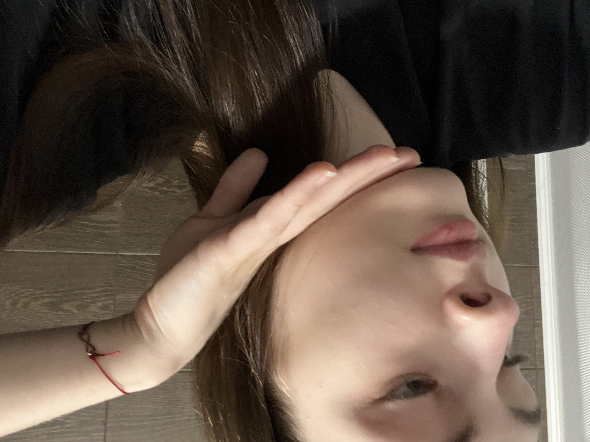 A close-up, indoor portrait captures the upper body of a young female, facing slightly left with her gaze directed downwards. Her fair-skinned face features light mascara on the eyelashes, natural eyebrows, and light pink lips. Long, dark brown hair falls over her right shoulder and chest. Her right hand supports her chin, with extended fingers resting along the jawline. A red string bracelet adorned with a small dark bead is visible on her right wrist. She wears a black garment, likely a shirt. The background to the left shows a vertical white, ribbed or textured surface, while the right features dark brown horizontal paneling with a wood grain texture. The image provides no visual cues to identify its specific location within the city of Saint Petersburg, Russia.