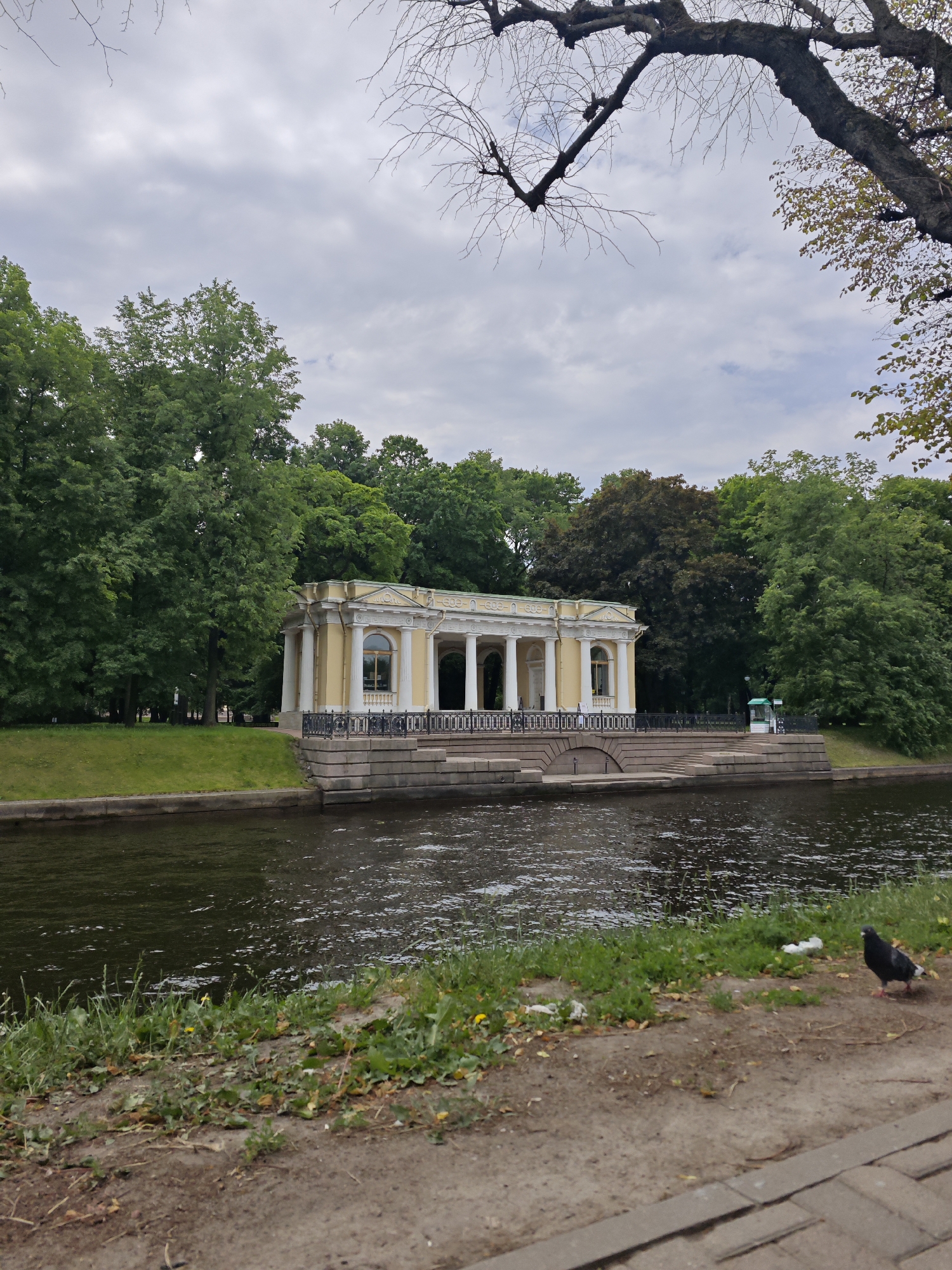 The image depicts a view of the Grotto Pavilion in the Mikhailovsky Garden, Saint Petersburg, Russia, on an overcast day. The pavilion, a neoclassical structure with a pale yellow facade and white columns, is situated on the bank of a dark, reflective canal. Lush green trees form a dense backdrop behind the pavilion, creating a serene, verdant atmosphere. The water in the canal shows ripples and a few scattered pieces of litter on the grassy bank in the foreground. A single pigeon stands on the pathway in the lower right corner of the frame, adding a touch of life to the scene. The overall impression is one of a quiet, picturesque park setting within the city.