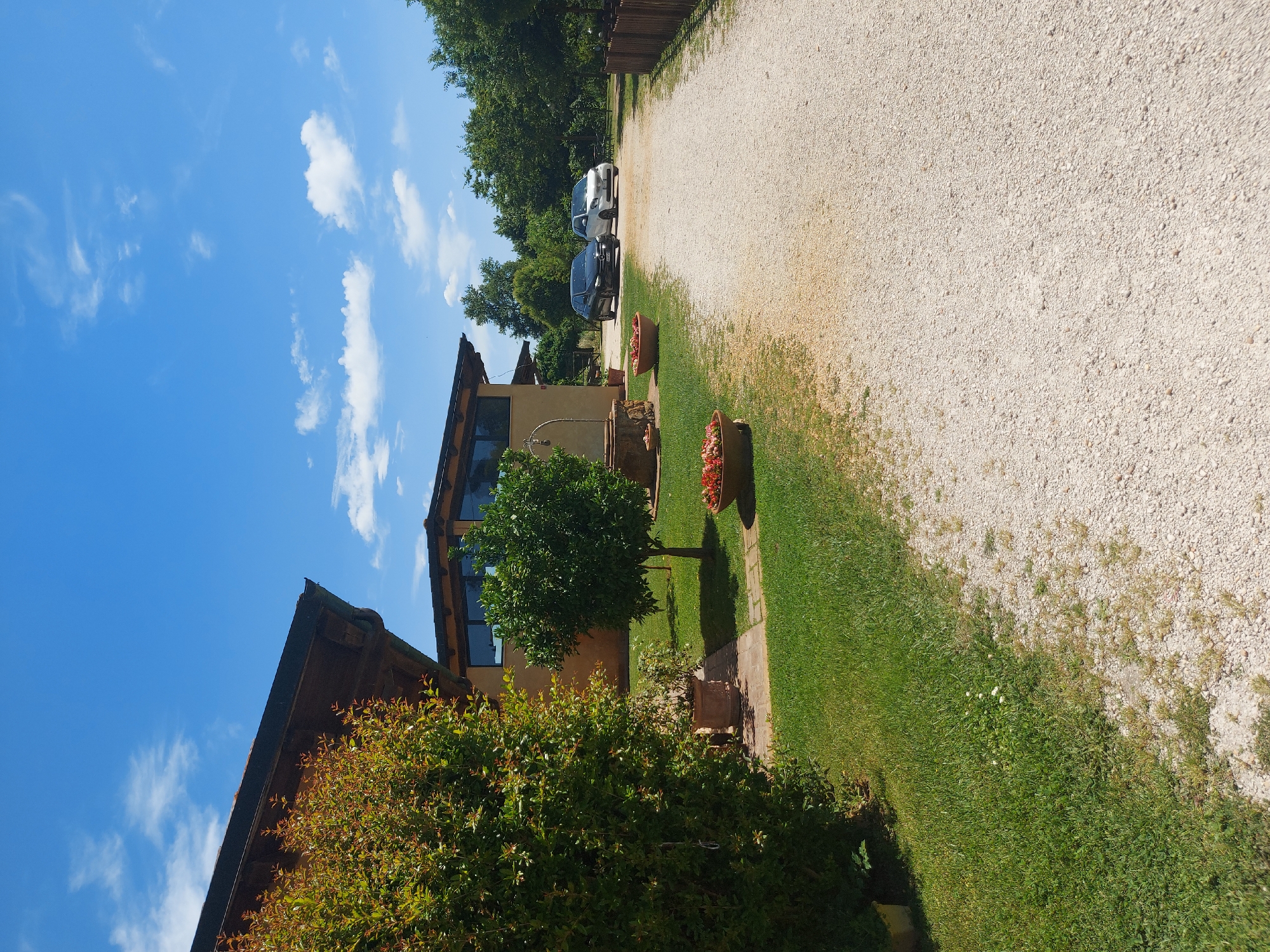 The image presents a daytime outdoor scene, likely in a rural or suburban area near Rome, Italy. A building with a modern design is visible, featuring a light-colored facade and large windows. Adjacent to the building is a gravel driveway leading to parked cars. The foreground includes well-maintained grassy areas, flower pots, and a small tree, suggesting landscaping. The sky is blue with scattered clouds. The photo is taken at an angle, resulting in a somewhat skewed perspective.
