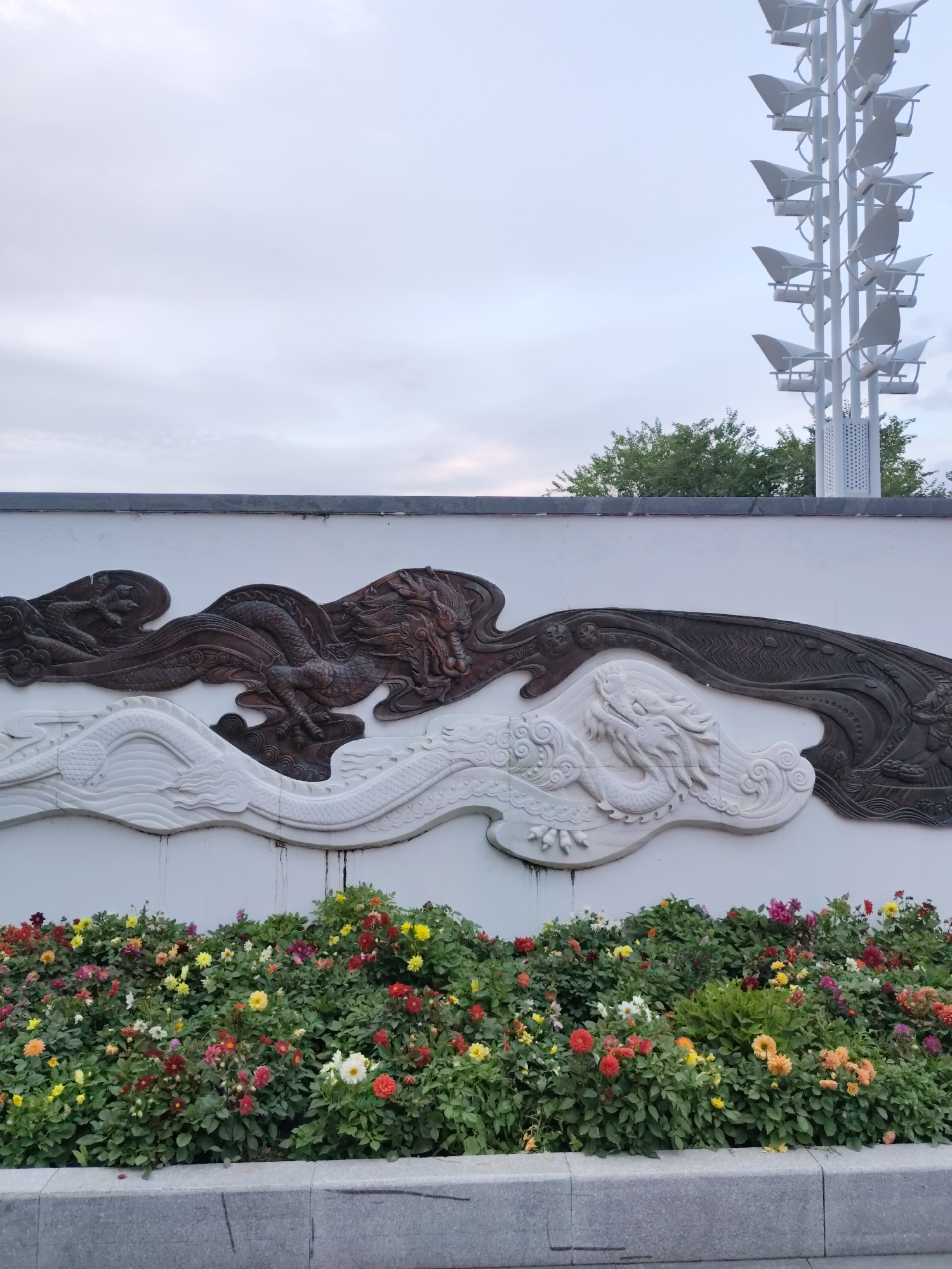 A low-angle, full outdoor shot captures a decorative wall in 幸福乡, China, adorned with intricate dragon carvings. The wall, painted white, features a relief sculpture of two dragons, one in a dark, textured material and the other in white, appearing to interact with swirling cloud motifs. Below the wall, a vibrant flowerbed overflows with a variety of colorful blossoms, including red, yellow, orange, pink, and white flowers, densely packed amidst lush green foliage. A grey, concrete border separates the flowerbed from a paved area in the foreground. In the background, a tall, white, modern sculpture with multiple light fixtures stands against a pale, overcast sky. Green trees are visible behind the wall and to the left of the sculpture. No people are present in the image.