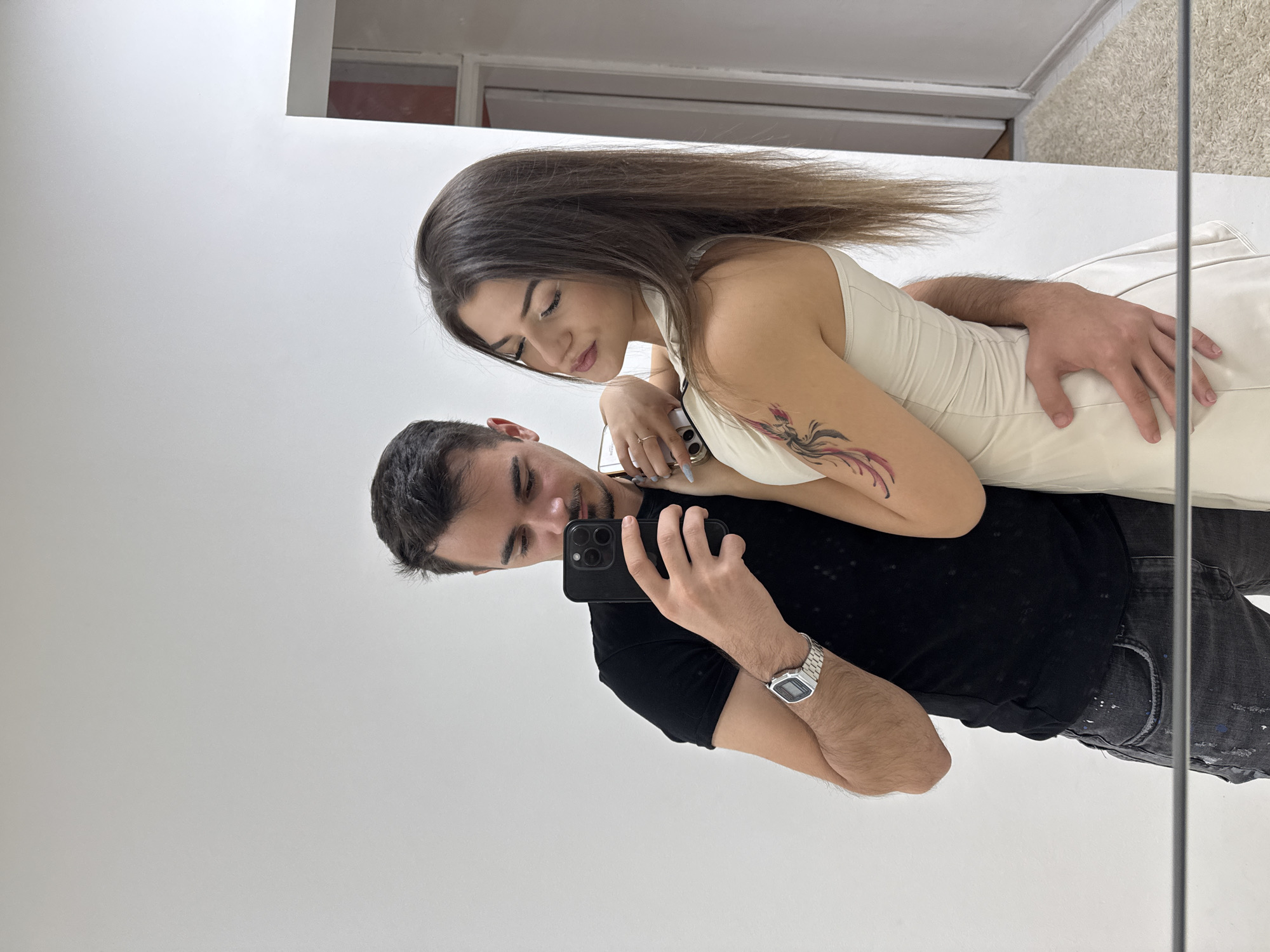 A man and a woman are taking a mirror selfie in what appears to be a white-walled room. The man, on the left, is wearing a black t-shirt and dark jeans. He has a watch on his left wrist and is holding a black smartphone to take the photo. The woman, on the right, is wearing a cream-colored dress and has a tattoo on her left arm. The man's right arm is around her waist. The background is a plain white wall, with a sliver of a doorway and a rug visible to the right. The scene is static and appears to be an indoor, domestic setting.