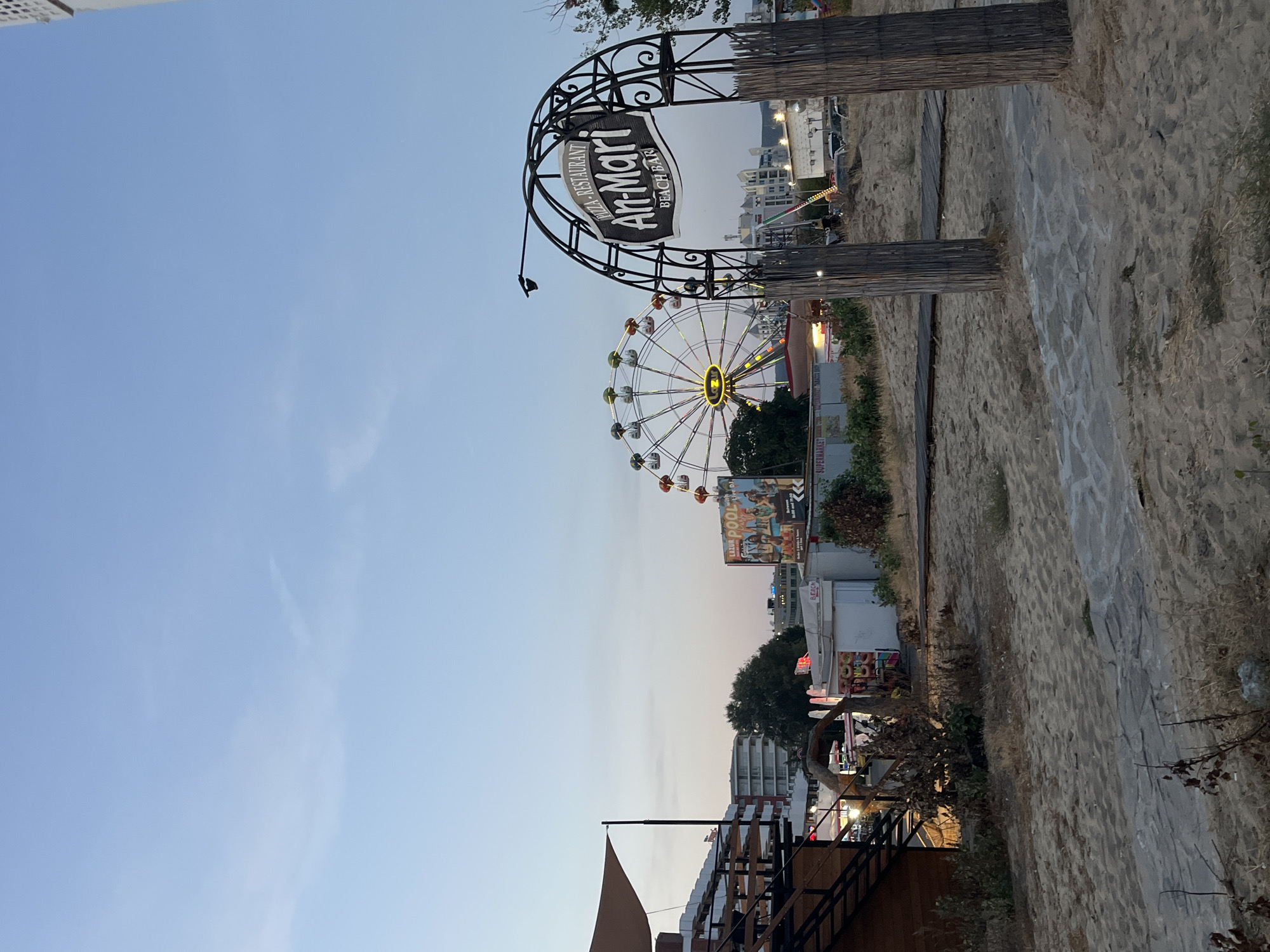 The image depicts a beach bar and amusement area in Nesebar, Bulgaria, at dusk. In the foreground, a sandy path leads through sparse vegetation towards a decorative arched gateway made of wrought iron and rustic wooden posts. The gateway is adorned with a sign reading "PIZZA-RESTAURANT An-Mari BEACH BAR."
Beyond the gateway, the scene opens up to a wider view of the beach area. To the left, a wooden structure with multiple levels and stairs, possibly a restaurant or viewing platform, is visible. Further back, a large Ferris wheel with illuminated cabins stands prominently against the pale sky, suggesting an amusement park or fairground. Beside the Ferris wheel, a billboard advertises "LAZUR POOL BAR" with images of people and drinks.
In the middle ground, several small kiosks and buildings are present, including one with a sign for "SUPERMARKET." The overall environment is sandy, with scattered dry vegetation and wooden boardwalks. Buildings and trees are visible in the background, and cars are parked in a lot further into the distance. The sky is a clear, soft blue, transitioning to a lighter shade near the horizon, indicating the late evening or early dawn. No people are clearly visible in the image.