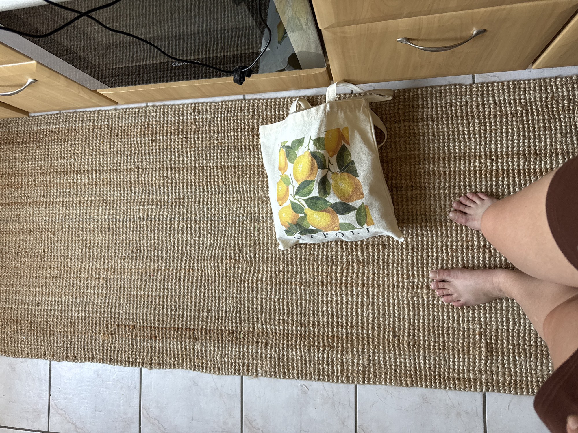 The image depicts a tightly woven, natural fiber rug on a tiled floor. The rug is a light brown, textured material. On the rug lies a white canvas tote bag with a print of lemons and green leaves. To the right of the rug, a wooden cabinet with a silver handle is visible, adjacent to a dark reflective surface, possibly a microwave or oven door. The lower portion of the image shows a person's bare feet and legs, indicating they are standing on the rug. The scene is indoors, in what appears to be a domestic setting. The image does not provide any specific visual cues to pinpoint a location within Höhenkirchen-Siegertsbrunn, Germany.