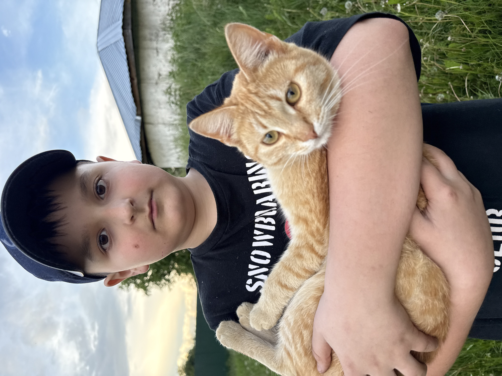 A young male child, approximately pre-teen, is depicted outdoors in Алегазово, Russia, holding an orange tabby cat. The boy, positioned centrally and looking directly at the viewer with a neutral expression, wears a dark blue baseball cap and a black short-sleeved t-shirt with partial white lettering visible ("SNOWBOAR"). A small dark mole is present on his right cheek (viewer's left). He cradles the ginger-colored cat horizontally across his forearms. The cat has yellowish-green eyes, a white muzzle, and white whiskers, and is also looking towards the camera with a relaxed posture. The background features a light-colored, single-story building with a peaked, corrugated silver metal roof on the right. The ground is covered in lush green grass and various wild plants, including some white seed heads. A blurry deciduous tree is visible to the left of the boy. The sky is overcast with light grey and blue clouds, indicating diffused natural light.
