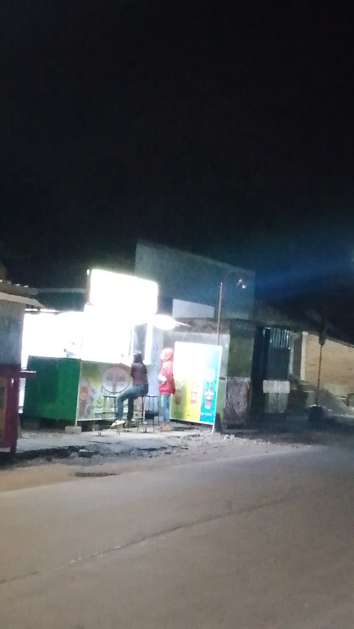 The image depicts a nighttime scene in front of a food stall or small shop on a paved street in Karanglangu, Indonesia. Two individuals are visible. One person, wearing a brown jacket and jeans, is seated on a stool at a counter. Another person, wearing a red hooded jacket and blue jeans, stands to the right of the seated individual. Behind them, the stall has a green base and a large, brightly lit white sign. To the right of the stall, there is a colorful poster or advertisement. The street in the foreground appears to be asphalt with some damage and debris. The background is dark, suggesting nighttime, with some ambient light from the stall illuminating the immediate surroundings.