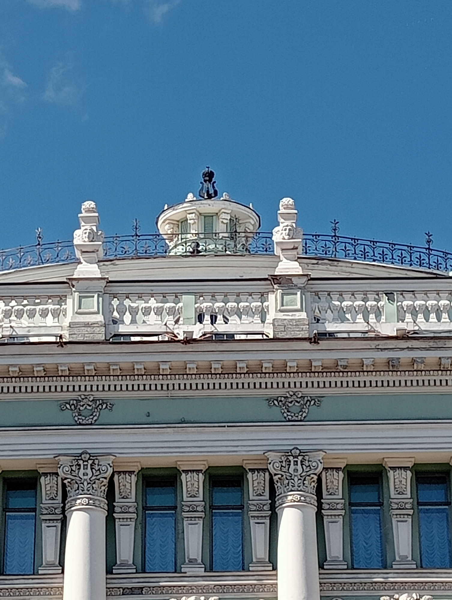 The image shows a close-up view of the upper facade of a building in Saint Petersburg, Russia, under a clear blue sky. The architectural style features ornate detailing, including decorative balustrades, sculpted figures, and columns.
Specifically, the facade exhibits a series of white, stone-like balustrades with repeating motifs. Above these, there is a dark decorative metal railing. Perched on pedestals along the roofline are carved stone elements, some appearing to be stylized faces or masks. At the very top center of the image, a dark, sculptural element, possibly an emblem or finial, is visible against the sky. The building's windows, visible in the lower portion of the frame, are rectangular and appear to be covered with blue curtains. The overall impression is one of classical or neoclassical architectural grandeur.