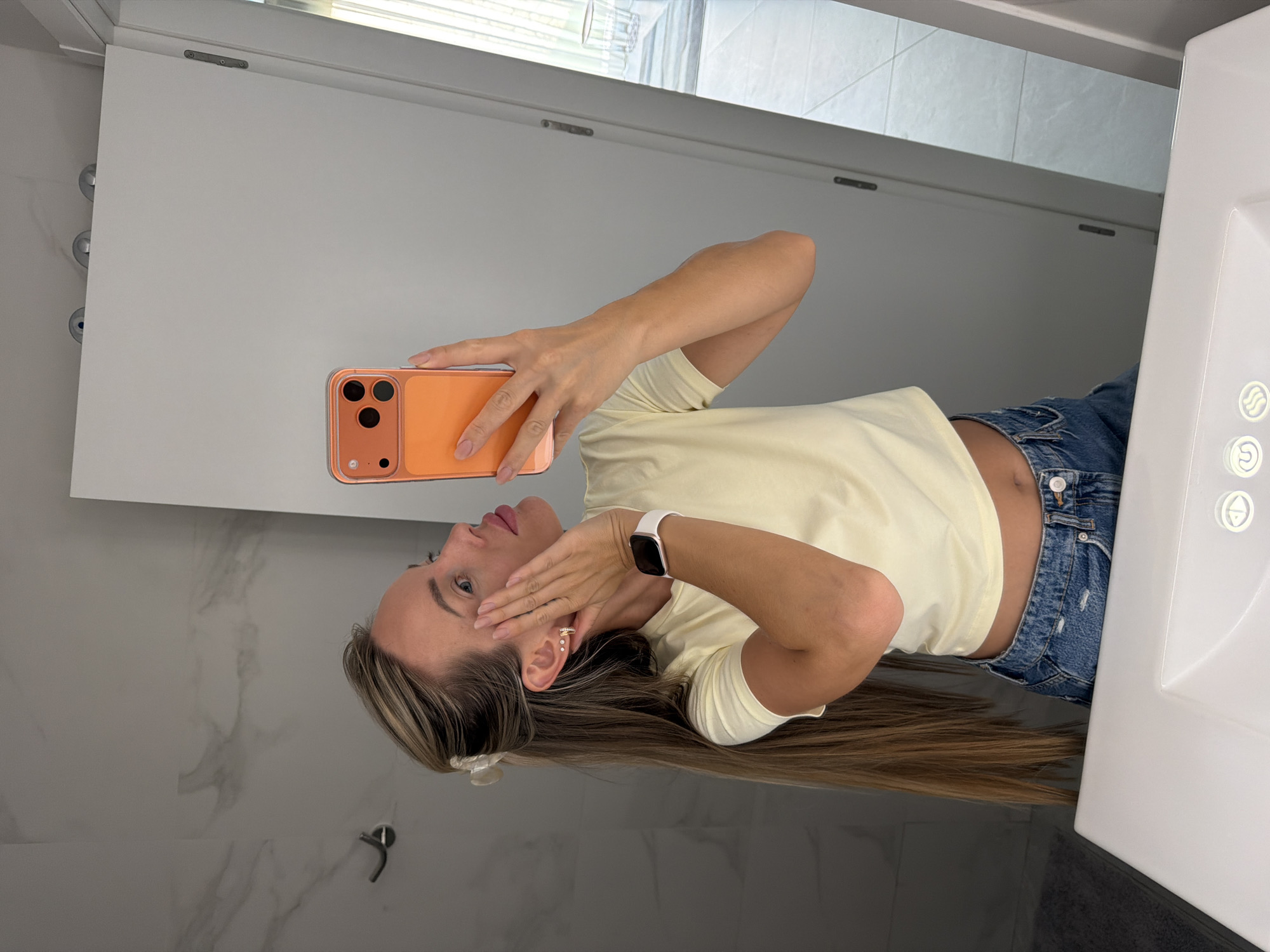 A woman is taking a mirror selfie in a modern bathroom. She is wearing a light yellow crop top and denim shorts. Her hair is long and blonde, partially pulled back with a clip. She is touching her face with her left hand and holding an orange smartphone in her right hand, which is angled to capture the reflection. Her right wrist has a white smartwatch. The bathroom features marble-patterned walls, a white sink, and a tall white door. The scene suggests a private setting, likely a home bathroom in Finestrat, Spain, given the context.