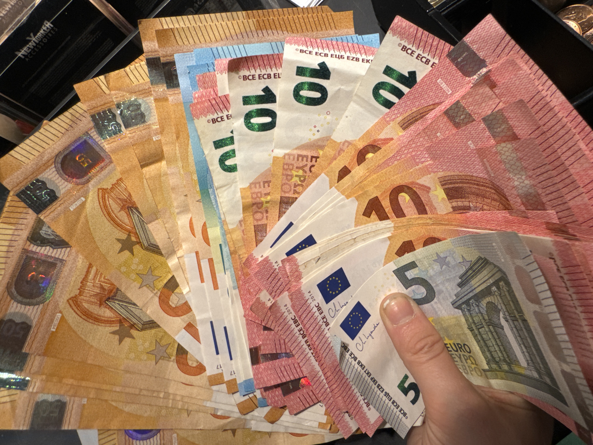 Here's a detailed description of the image:
**Subjects and Objects:**
* A hand holding a fan of Euro banknotes of various denominations: €5, €10, and €50.
* The banknotes are layered, with the €5 notes appearing more prominently in the foreground, followed by the €10 and €50 notes.
* On the side, there is a container containing a box "NEWYORKER ACCESSORIES" and coins.
**Actions and Interactions:**
* A hand is holding the fan of Euro banknotes.
* The banknotes are oriented to show their face values.
**Environment:**
* The background is mostly dark, suggesting an indoor setting with low lighting.
* The composition focuses on the banknotes, with the surrounding environment providing context.
There is no indication of the location being within the city of Unknown, Unknown.