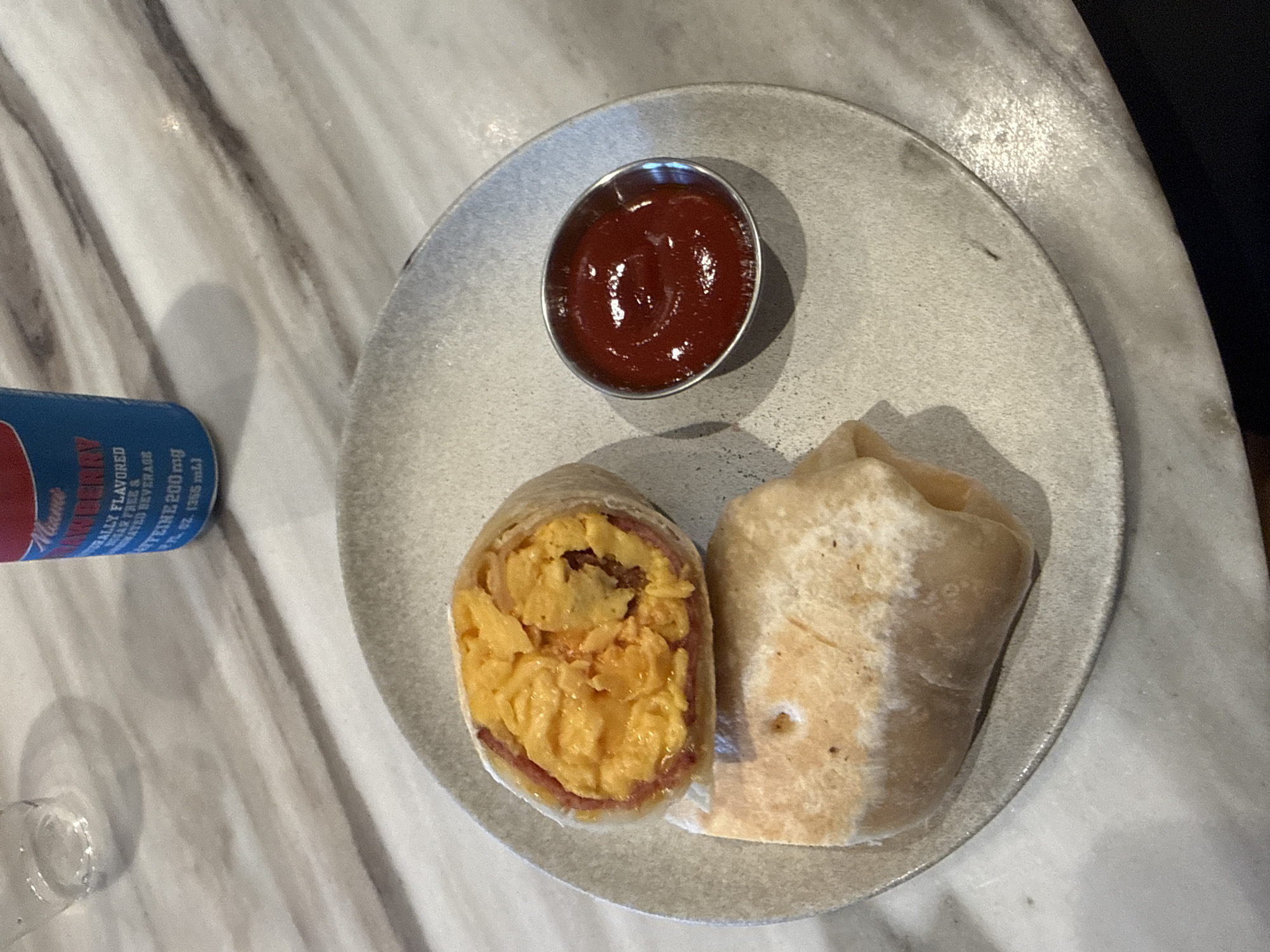 The image shows a plate with a breakfast burrito cut in half, accompanied by a small metal container of ketchup. The burrito appears to contain scrambled eggs and ham. The plate is placed on a marble-patterned surface next to a partially visible blue can. The scene is likely indoors at a restaurant or cafe, potentially in Los Angeles, based on the general food presentation and drink choice that are common in the area.