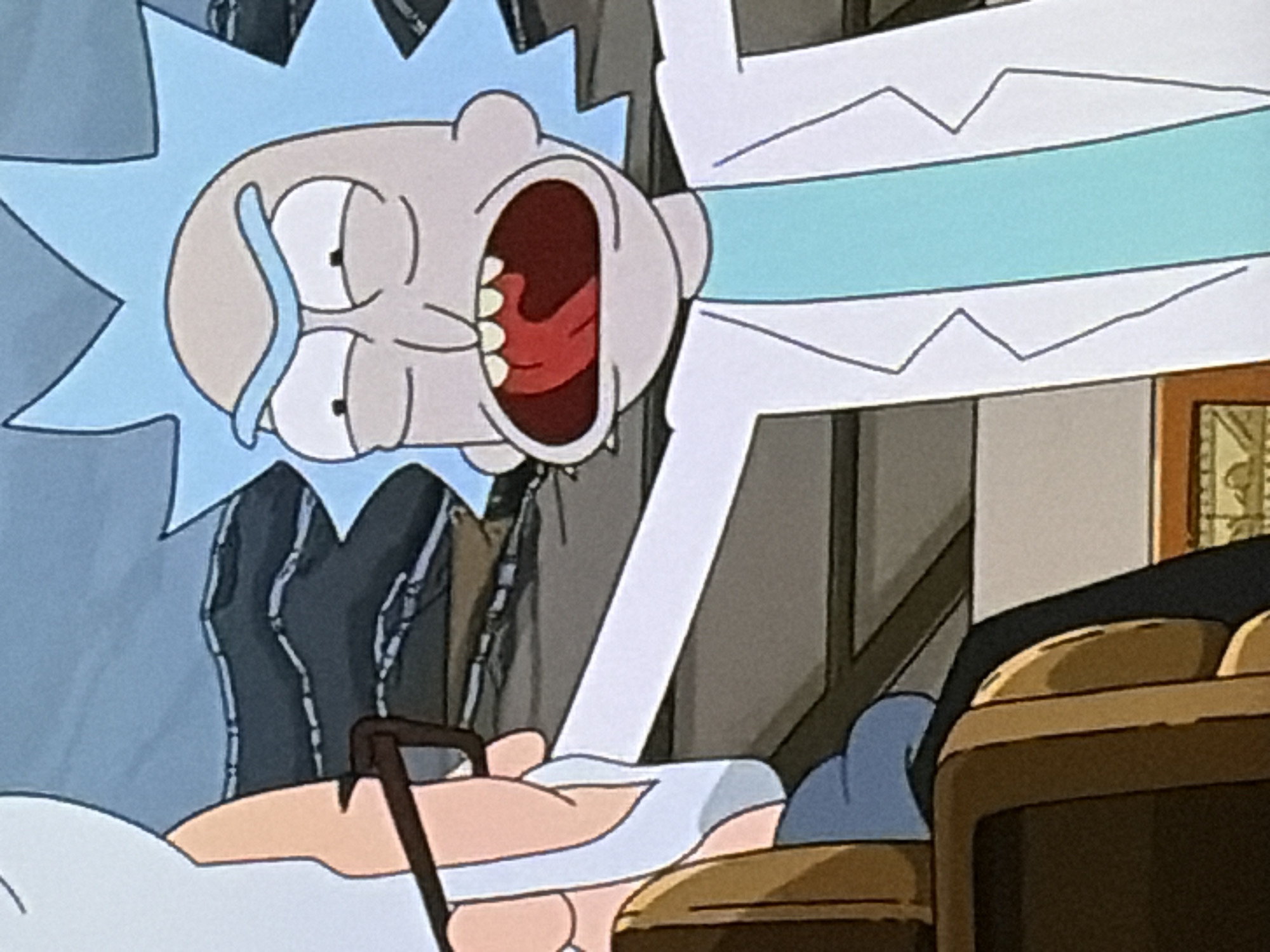 In a medium shot, two animated characters are depicted. On the left, an elderly man wearing glasses and a white beard is partially visible, seated in a brown chair with a blue garment. On the right, a man with light blue spiky hair, wearing a lab coat over a turquoise shirt, is facing the elderly man with his mouth wide open, revealing his tongue and teeth. The background consists of a dark, layered surface, possibly representing a landscape or a stage. The scene appears to be from an animated show or film.