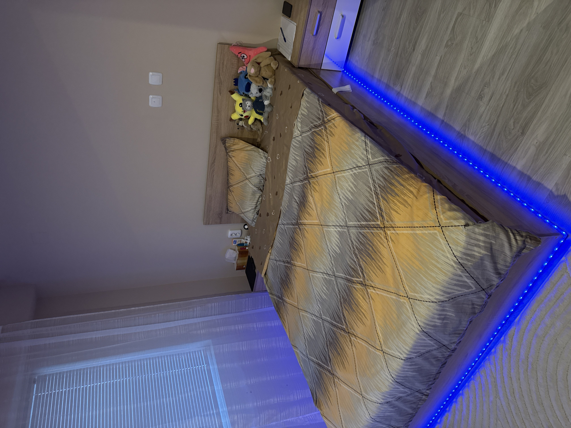 A bedroom scene photographed indoors in Lovech, Bulgaria, features a bed with a gray and yellow patterned duvet cover, and a headboard with plush toys. A nightstand is positioned next to the bed, illuminated by blue LED strip lights which extend along the edges of the bed. The wall features light switches. A window with blinds is visible. The overall lighting in the room is a mix of natural and artificial, influenced by the LED lights.