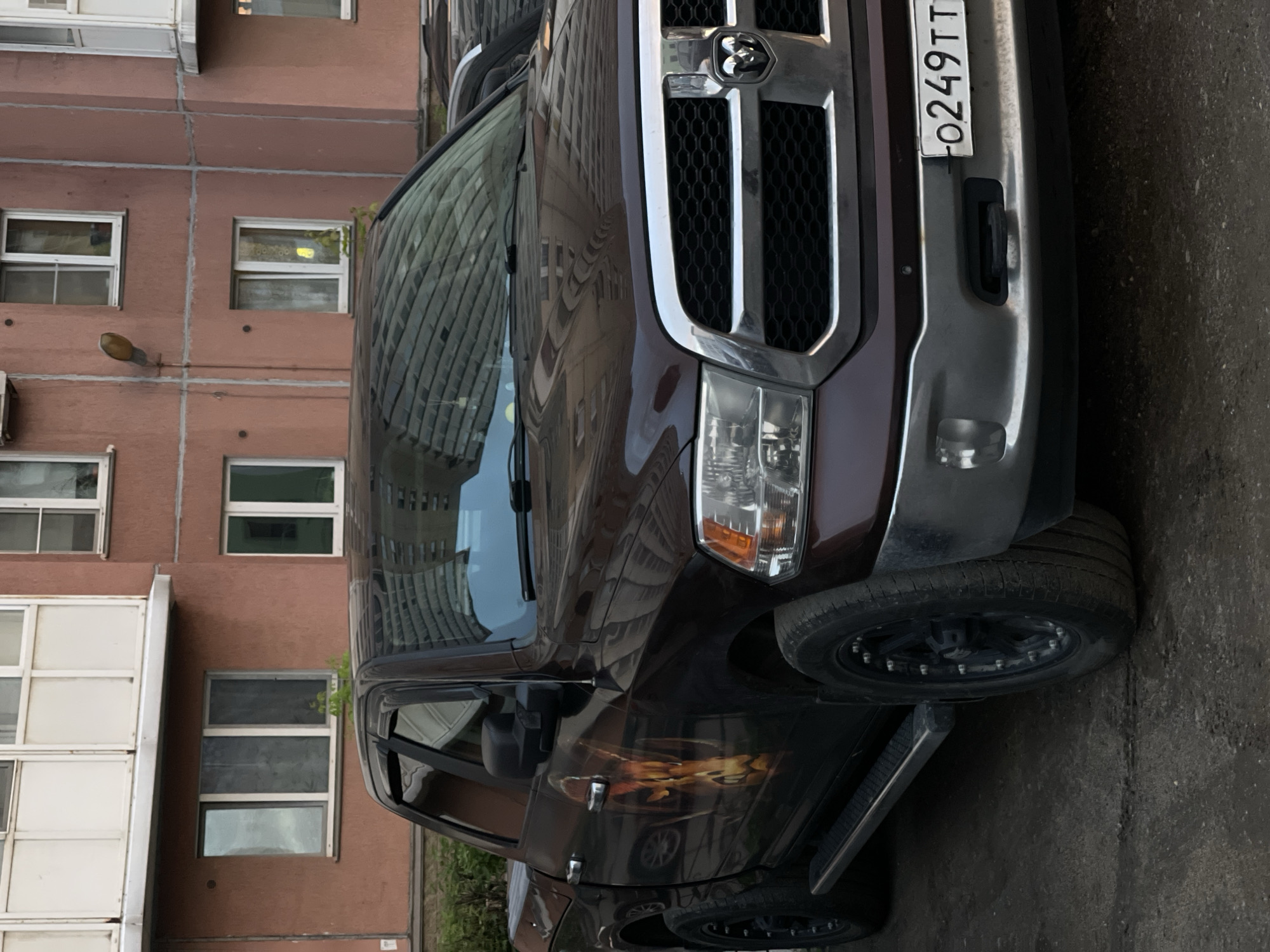 A dark red Ram pickup truck with a graphic of a ram's head on the driver's side door is parked on a paved surface in front of a multi-story building. The truck has a chrome grille and bumper. The building has rows of windows and a light-colored trim. The sky is reflected in the truck's windshield.