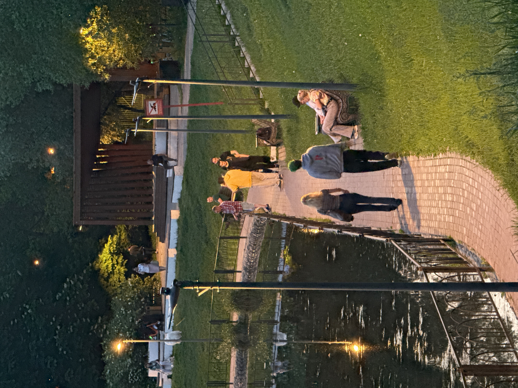 Here is a description of the image.
The image shows an outdoor scene at night, likely in a park-like setting in Moscow, Russia. A paved walkway winds along a body of water, possibly a canal or small lake. Along the walkway, there is a wrought-iron fence. Several people are visible along the path, some standing and talking, others sitting on benches. The area is illuminated by streetlights. The lighting suggests it is dusk or night. Lush greenery, including trees and grass, surrounds the walkway and body of water. There are various park features, such as a fence or structure with a sign, further in the background.