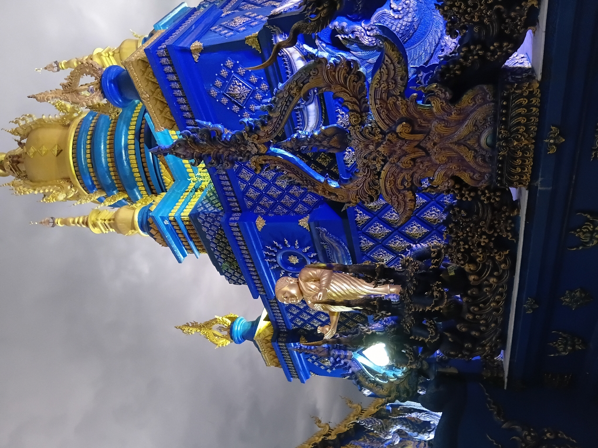 The image shows a detail of the Wat Rong Suea Ten, also known as the Blue Temple, in Chiang Rai City Municipality, Thailand. The temple is characterized by its striking sapphire blue exterior, adorned with intricate golden embellishments. In the foreground, a golden statue of a standing Buddha is positioned amidst ornate, dark, sculpted figures and architectural elements. Behind the statue, the blue and gold tiered structure of the temple rises, with its layered roofs and decorative spires visible against a cloudy sky. The lighting suggests it is either dusk or nighttime, with artificial lights illuminating the temple's features. The architectural style is distinctly Thai, featuring elements like multi-tiered roofs and ornate carvings.