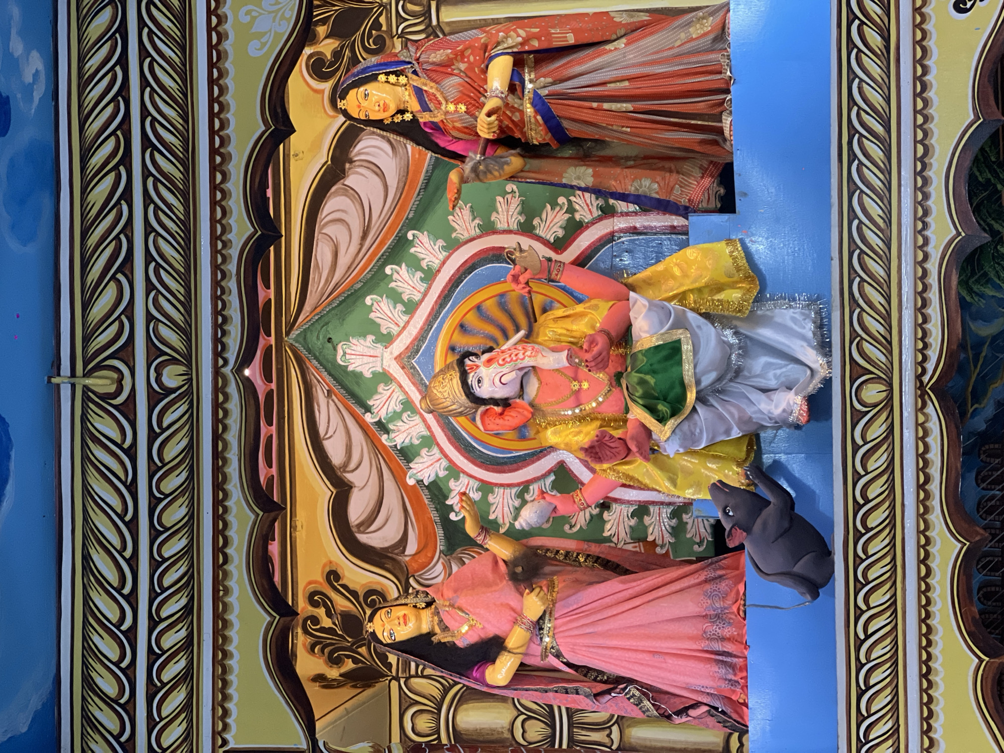 This image shows a painted mural or tableau likely located within a temple or religious building in Varanasi, India. The central figure is a depiction of the Hindu deity Ganesha, recognizable by his elephant head. He is painted with a pink body, yellow cloth, and white lower garment. He holds a conch shell in one hand and a club in another. Flanking Ganesha are two female figures, one above him in a red and orange sari, and one below in a pink sari. The tableau has an elaborate painted background featuring green, white, and beige tones, surrounded by decorative borders. A gray rat, Ganesha's vahana (vehicle), is depicted near the bottom figure. The entire scene is set against a blue backdrop.