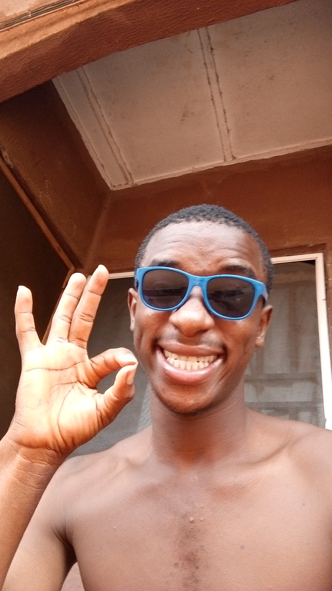 A young Black man is captured in a close-up, smiling broadly at the camera. He wears blue-framed sunglasses that cover his eyes. His wide grin reveals his upper and lower teeth, and subtle dimples are visible on his cheeks. He is shirtless, displaying his shoulders and upper chest. His left hand is raised to the left of his face, making an "OK" gesture with his thumb and forefinger forming a circle, while the other three fingers are extended upwards.
The background suggests an indoor or semi-enclosed environment, possibly a porch or a room with an open structure, typical of residential settings in places like Onitsha, Nigeria. Warm brown, possibly wooden, architectural elements are visible at the top and sides, resembling ceiling beams or door frames. Behind the man's head, a lighter-colored ceiling or upper wall section with faint lines or texture is present. A white window or doorway frame with a dark mesh screen is partially visible to the right. The bright lighting indicates it is daytime. The man's expression conveys happiness, cheerfulness, and confidence. No visible text is present in the image.