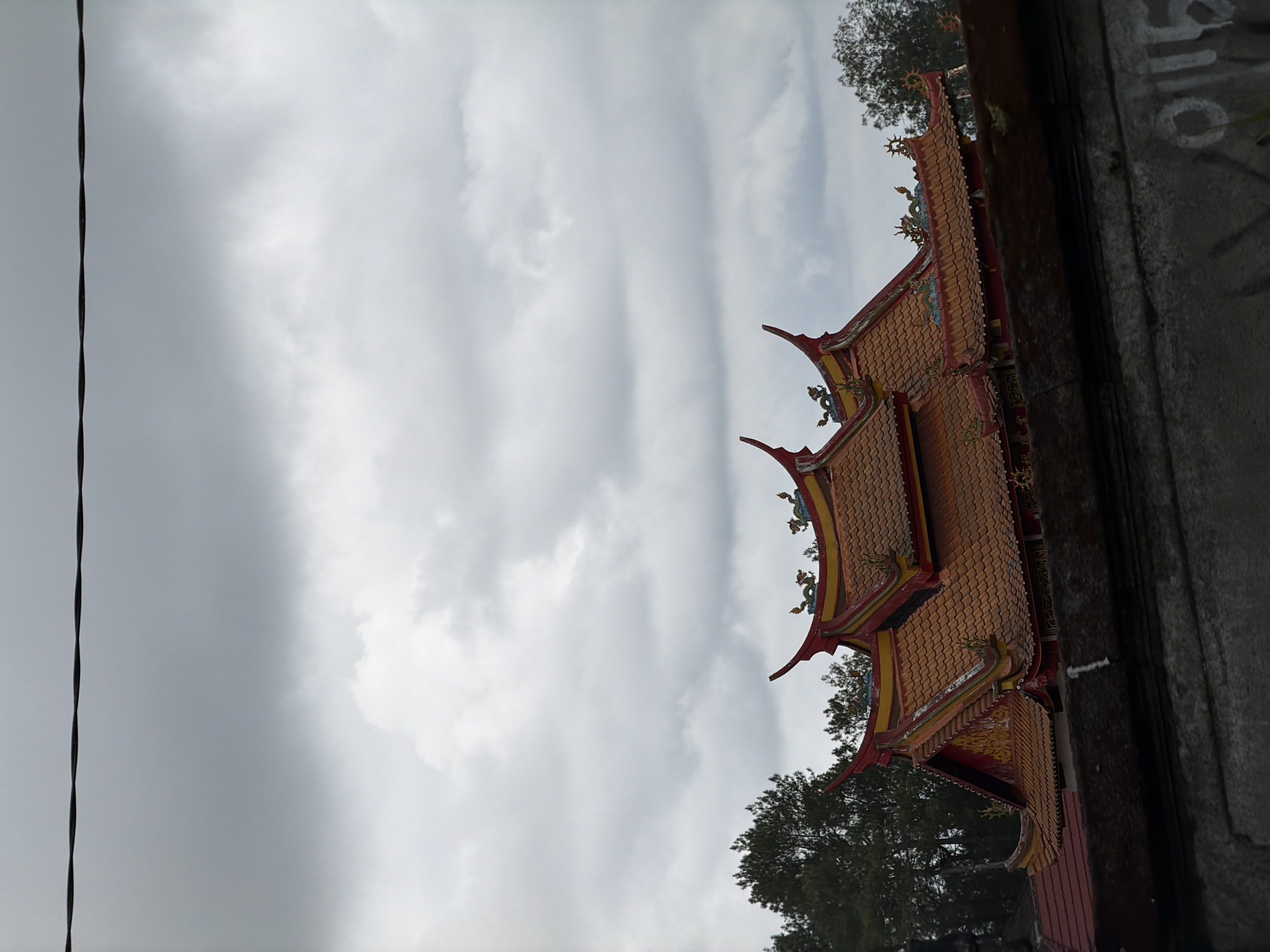 In Kintamani, Indonesia, a traditional temple building with orange, tiled roofs and ornate red and gold decorations stands against a backdrop of a cloudy, overcast sky. The temple features distinct architectural elements, including curved rooflines, pointed finials, and decorative dragon sculptures along the ridges. Dense green trees are visible to the left of the temple, contrasting with the muted tones of the sky. In the foreground, a dark, textured structure, possibly a wall or railing, partially obscures the view of the temple's lower sections. A power line stretches horizontally across the upper portion of the frame. The overall atmosphere is somber due to the cloudy weather.