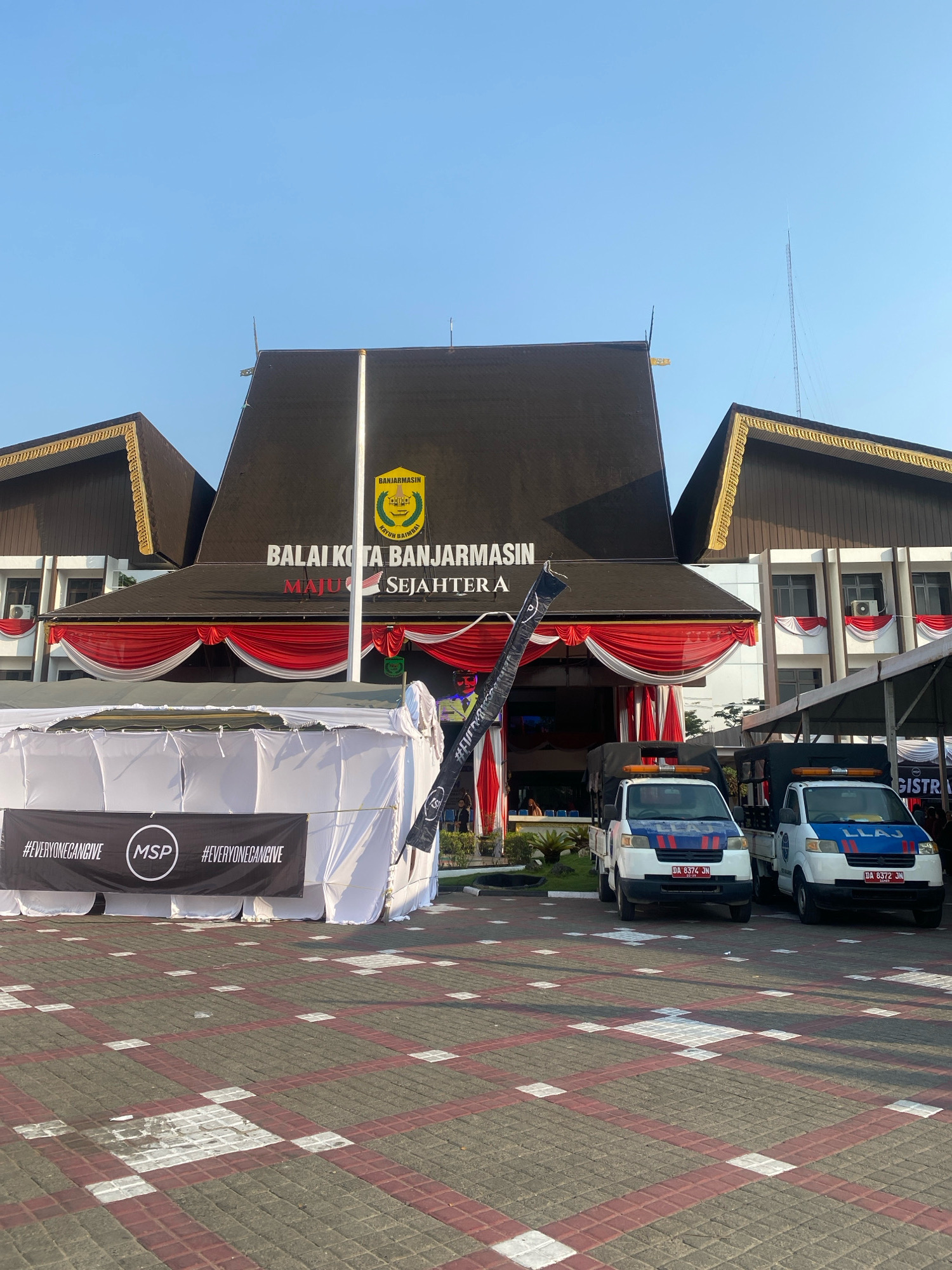 This is a photograph taken outdoors, likely in the city of Banjarmasin, Indonesia, in front of a government building identified as "BALAI KOTA BANJARMASIN" (Banjarmasin City Hall). The building features a distinct dark, pitched roof with ornate golden trim along the eaves. Red and white drapery is hung across the facade of the building, suggesting a celebratory or official event.
In the foreground, two white pickup trucks with blue and red decals are parked on a paved area. The truck on the left has the license plate "DA 8374 JN," and the truck on the right has the license plate "DA 8372 JN." The truck on the right also displays the text "LLAJ." In front of the trucks, a white temporary structure or tent is erected, with a black banner across it. The banner contains the text "#EVERYONEGIVE" on either side of a circular logo with "MSP" inside. A black flag with a logo is also visible to the right of the banner. The ground is paved with interlocking bricks in a pattern of gray, red, and white squares and rectangles. The sky above is clear and blue, indicating a daytime scene, likely morning or afternoon. The overall impression is one of an organized outdoor event or gathering at a significant civic location.
