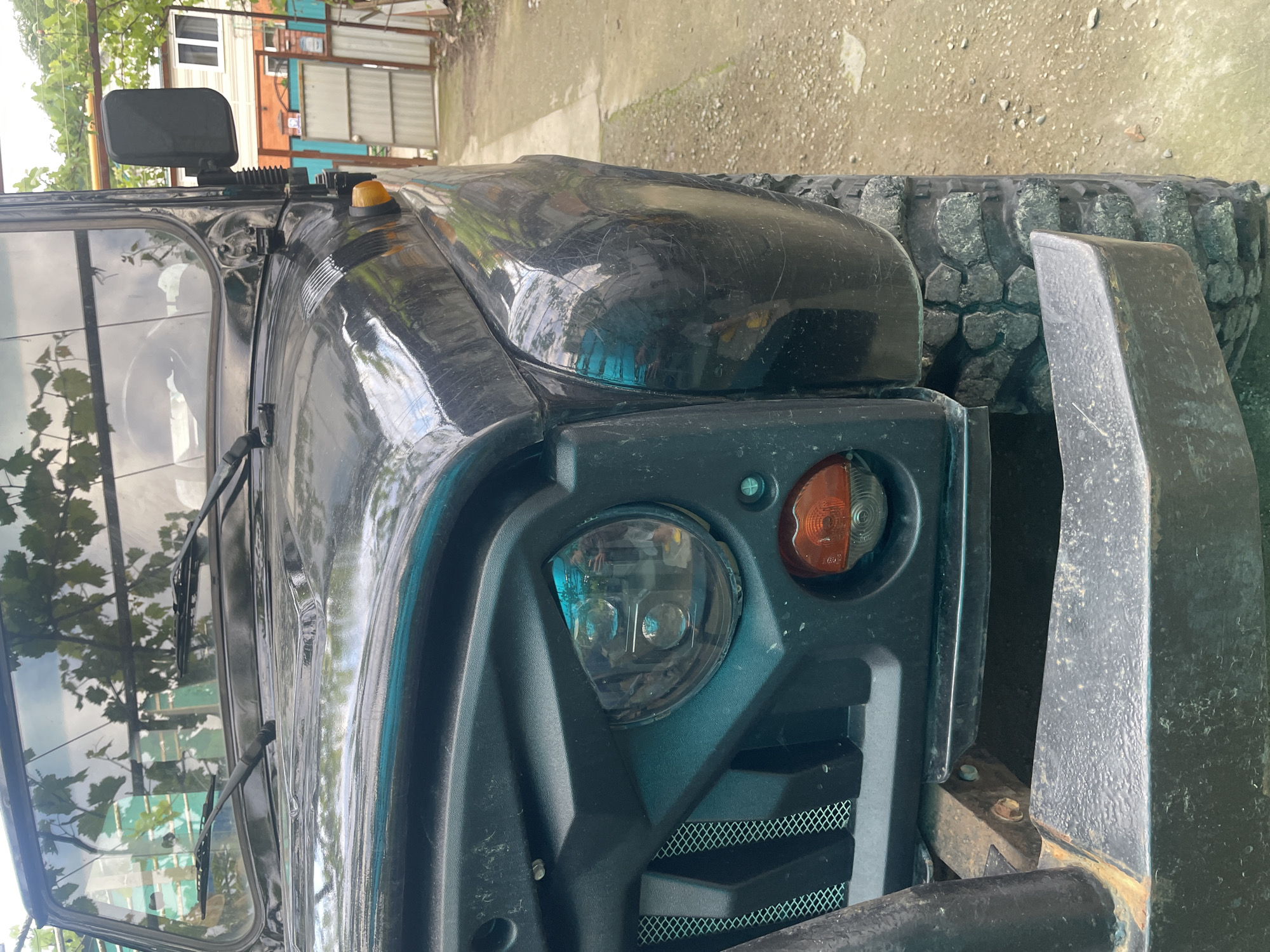 The image shows a close-up view of the front of a black, rugged vehicle, possibly a modified SUV or off-road vehicle. Prominent features include the headlight, turn signal, front bumper, and a portion of the tire. The windshield and a side mirror are visible. The background appears to be an outdoor environment, perhaps a residential area. The quality of the vehicle's paint suggests it has been used. There are signs of foliage and a building partially visible in the background. The image does not provide conclusive evidence of being in Olginka, Russia.
