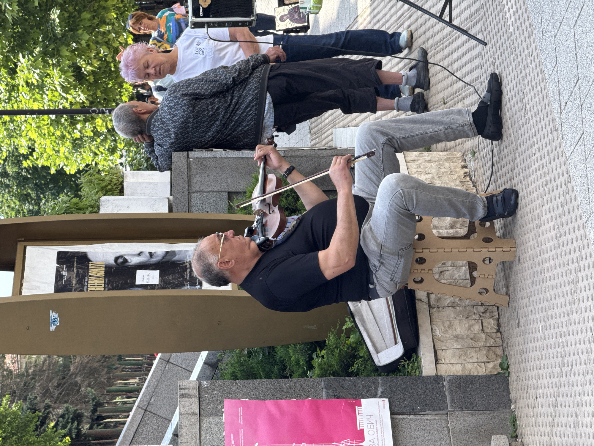 A street performer plays the violin on a fold-up stool in front of a building in Pleven, Bulgaria. He is a man with grey hair and glasses, wearing a black t-shirt and grey jeans. To his right, a group of people stand and observe. One woman with pink hair wears a white t-shirt with a Loewe logo. A man with grey hair leans on a ledge. A woman with brown hair stands in the background wearing a colorful shirt. Posters are visible on the building behind them, one a large black and white poster with a woman's face, and another a pink poster on the left. The ground is paved with grey tiles.