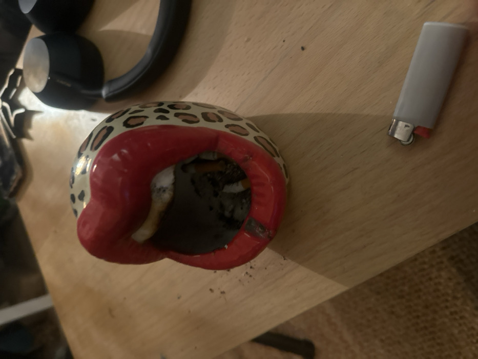 The scene depicts a close-up, elevated view of a light-toned wooden surface. A prominent ceramic ashtray, designed as a pair of open, glossy red lips against a cream-colored background with brown leopard print spots, occupies the center-left. Inside the ashtray, multiple cigarette butts are visible amidst a collection of dark ash. A distinct dark burn mark is present on the lower red lip rim of the ashtray, with scattered ash debris on the wooden surface surrounding it. To the lower right of the ashtray, a gray disposable lighter with a red ignition button rests on the wooden surface. In the upper right background, a pair of dark, possibly black, over-ear headphones is partially visible, appearing out of focus. Other indistinct objects are blurred in the far background. The environment appears to be an indoor setting under artificial lighting.