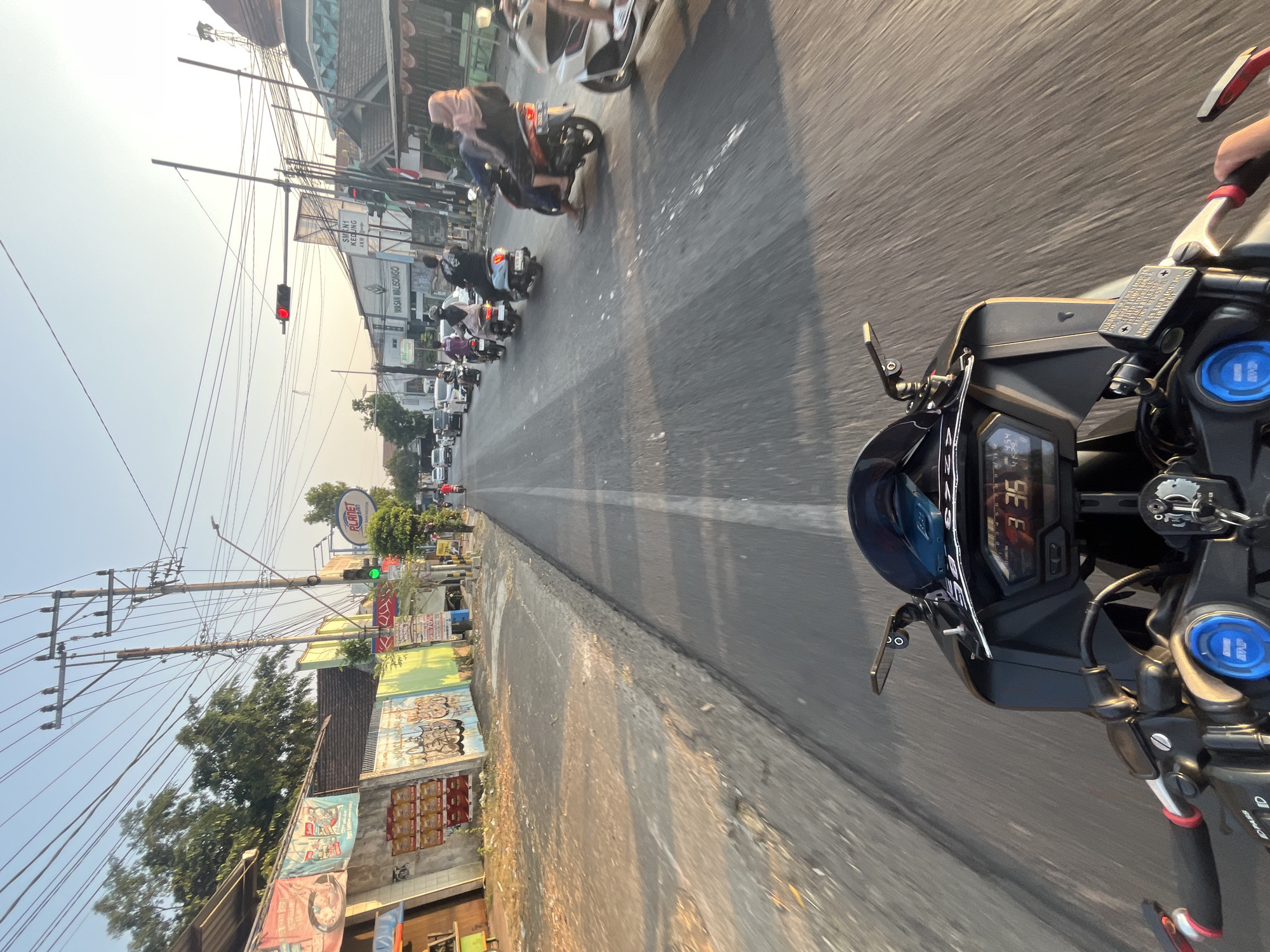 The image is a first-person perspective shot from a motorcycle on a street in Pecangaan, Indonesia. The road is paved with a white line dividing the lanes. On the left side of the road, there are buildings with signage, including advertisements and graffiti. A traffic light is visible in the distance, and it is red, indicating that traffic should stop. Several other motorcycles and a few cars are on the road, moving in the same direction as the motorcycle in the foreground. The sun is setting, casting long shadows and illuminating the scene with warm, golden light. The motorcycle's dashboard shows a speed of 33 km/h and the time 3:35. There are power lines and utility poles running along the side of the road. The environment appears to be a busy urban or semi-urban area.