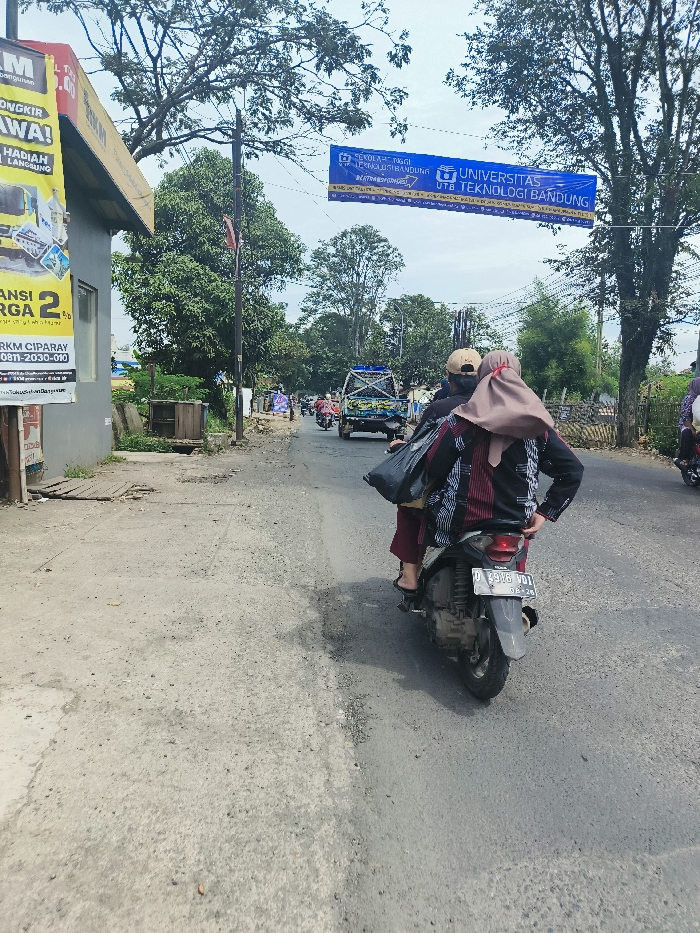 The scene is a street in Ciparay, Indonesia, located at the coordinates -7.027 latitude, 107.693 longitude. Two people are riding a black motorcycle on a paved road. The person in the back is wearing a brown hijab and a striped shirt. The person in the front is wearing a white shirt. They are both facing forward, and the motorcycle is moving to the right. The motorcycle is facing forward and is moving in the same direction as the people. The motorcycle has a black license plate on the rear, and it reads "D 2316 VDI". There are trees and buildings on both sides of the street. A blue banner above the street reads "Universitas Teknologi Bandung." There is a small truck in the distance with a blue light on top, and a person is walking on the right side of the street. The street is paved and the road is slightly gray. The weather is cloudy. There is a sign on the left side of the street. It is yellow and blue and has writing in Indonesian. The road is relatively empty, and there is no visible traffic. The scene is calm and tranquil.
