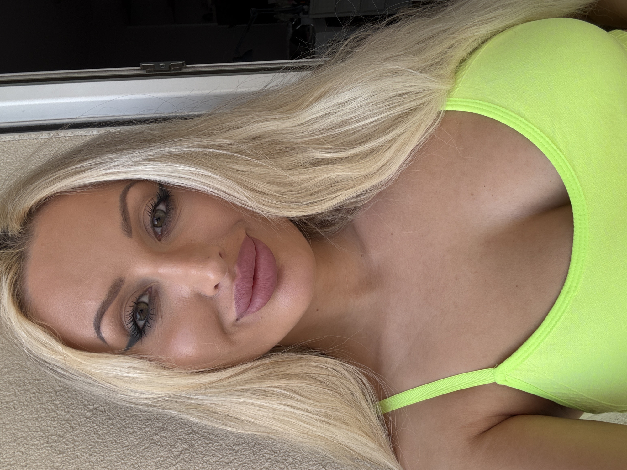 A close-up, frontal portrait features a light-skinned female with long, straight platinum blonde hair, styled to fall over her shoulders and upper chest. She has hazel eyes with black winged eyeliner, defined arched eyebrows, and full lips colored with a matte pink lipstick, exhibiting a slight smile. She is wearing a neon lime green top with a scoop neckline and thin shoulder straps, one visible on her left side. The background to the left consists of a textured, light beige wall. To the right, a white door frame with a visible metal hinge is partially open, revealing a dark interior space. The scene is located indoors in the city of Unknown, Unknown.