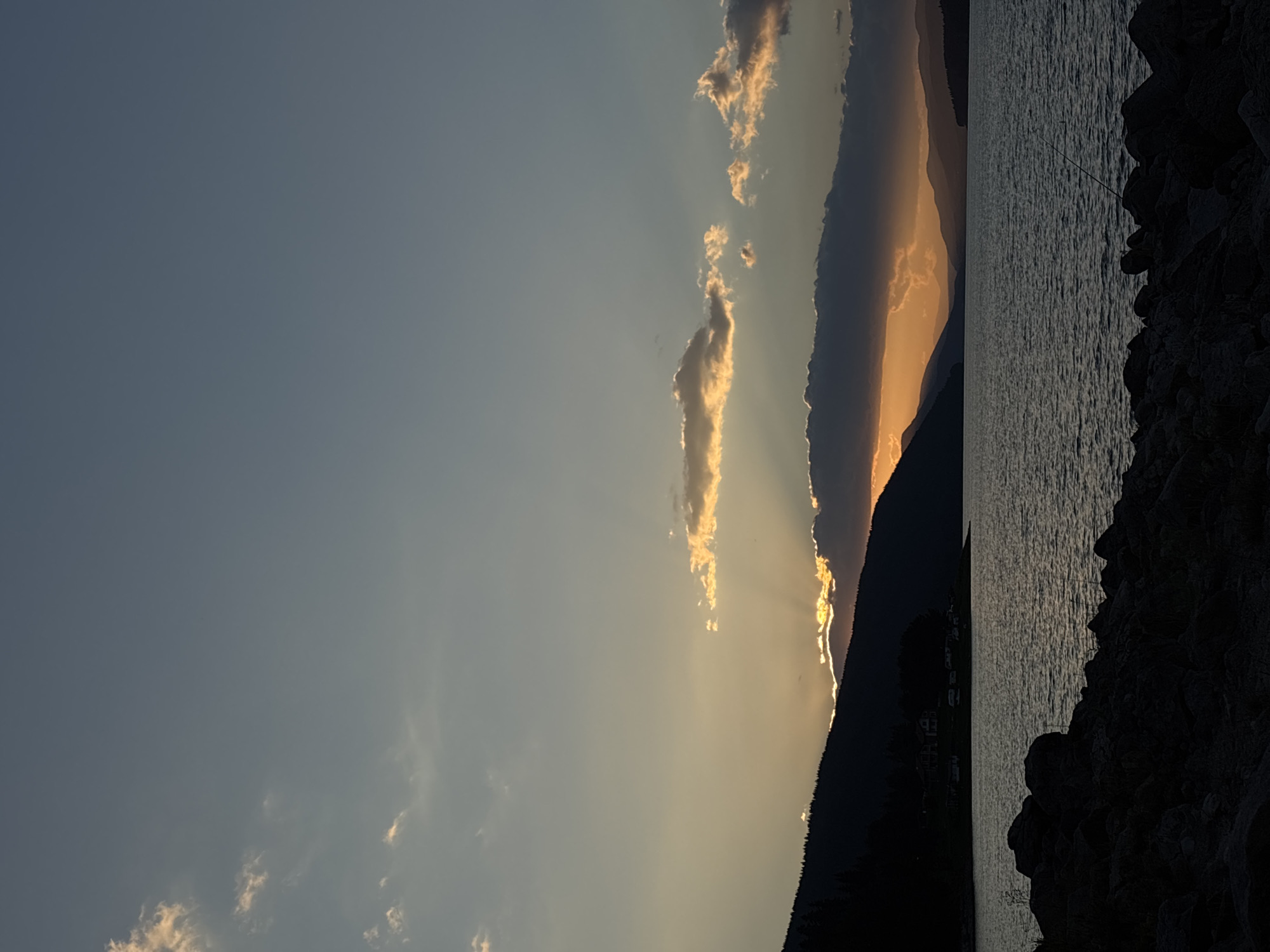 This is a vertical, low-angle, outdoor photograph taken at dusk. The scene depicts a body of water, likely a lake, with a silhouetted shoreline of hills and mountains in the background. The sky is a gradient of soft blues and grays, with scattered clouds illuminated by the setting sun. Sun rays are visible, bursting from behind a dark cloud formation that partially obscures the horizon. The water's surface is textured with ripples, reflecting the muted light. In the foreground, dark, rough rocks line the edge of the water. A single fishing rod is visible on the right side of the frame. A small settlement with buildings and a few campers is visible on the distant shoreline, nestled amongst trees. The overall atmosphere is serene and tranquil. The location is Unknown, Unknown.