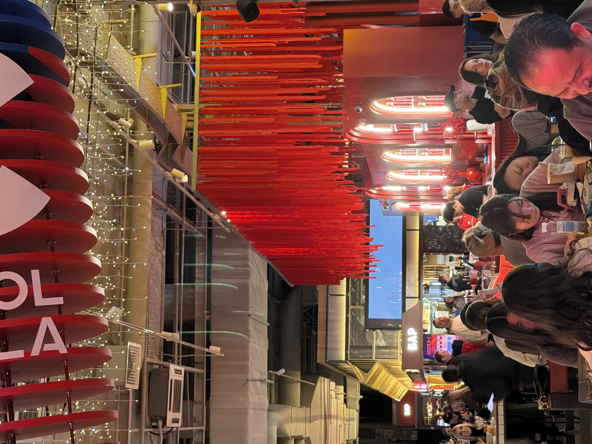The image depicts an outdoor urban scene in Moscow, Russia. The view is oriented lengthwise along a street featuring outdoor dining areas on both sides. Numerous people are seated at tables, suggesting a restaurant or cafe setting. Overhead, there are architectural decorations: a series of horizontal red elements (perhaps streamers or slats) and a red-and-white sign (possibly a "Coca-Cola" display) with lights. Building facades are visible, and the street is illuminated by various light sources. The overall impression is that of a busy, vibrant public space.