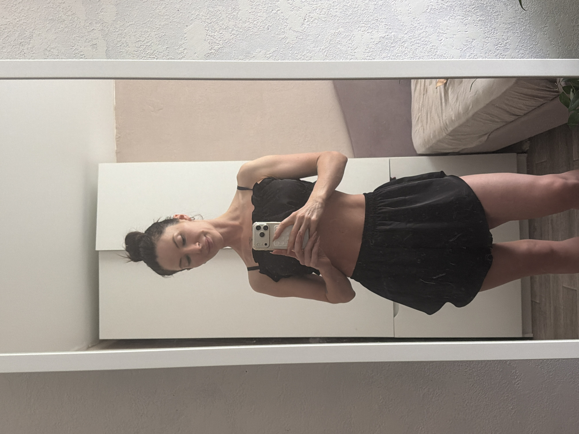 A young woman is depicted taking a mirror selfie in an indoor setting, likely a home in Perm, Russia. She is facing slightly towards the left of the frame, holding a smartphone with a white case in her right hand, which is angled across her torso. Her dark hair is styled in an updo or bun, and she has a subtle, closed-mouth smile, with a nose piercing visible. She is wearing a black bralette or camisole top and black shorts.
The mirror's reflection shows parts of the room behind her. A light-colored, textured wall is visible in the upper portion, along with a smoother, light-beige section. In the upper right of the reflection, a bed with light-colored (possibly tan or beige) bedding is partially seen. To the left, behind the woman, a white dresser or cabinet is reflected. A light-colored wooden floor is visible in the lower right, and a hint of a green plant appears near the bed. The mirror itself has a clean white frame. The scene conveys a relaxed, casual moment. No specific time of day or external weather is indicated by the indoor lighting. There is no visible text in the image.