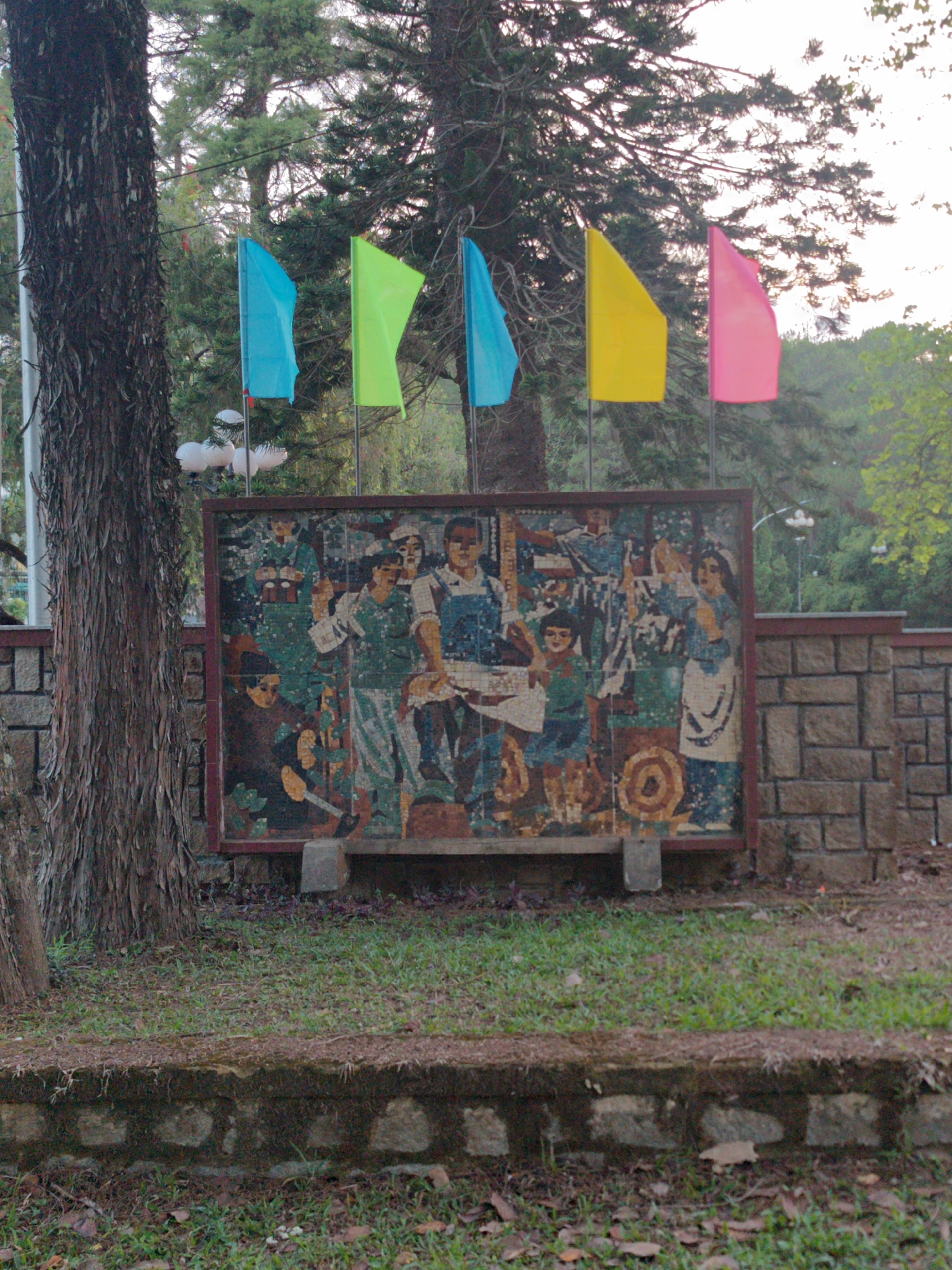 The image captures an outdoor scene in Cam Ly - Đà Lạt, Vietnam. Five colorful flags, blue, lime green, blue, yellow, and pink, are planted behind a large mosaic artwork depicting a group of people, likely laborers. The mosaic is placed in front of a stone wall. A tall, textured tree trunk stands to the left of the mosaic, and the ground is covered in grass and fallen leaves. Additional trees are visible in the background under a sky of even brightness.