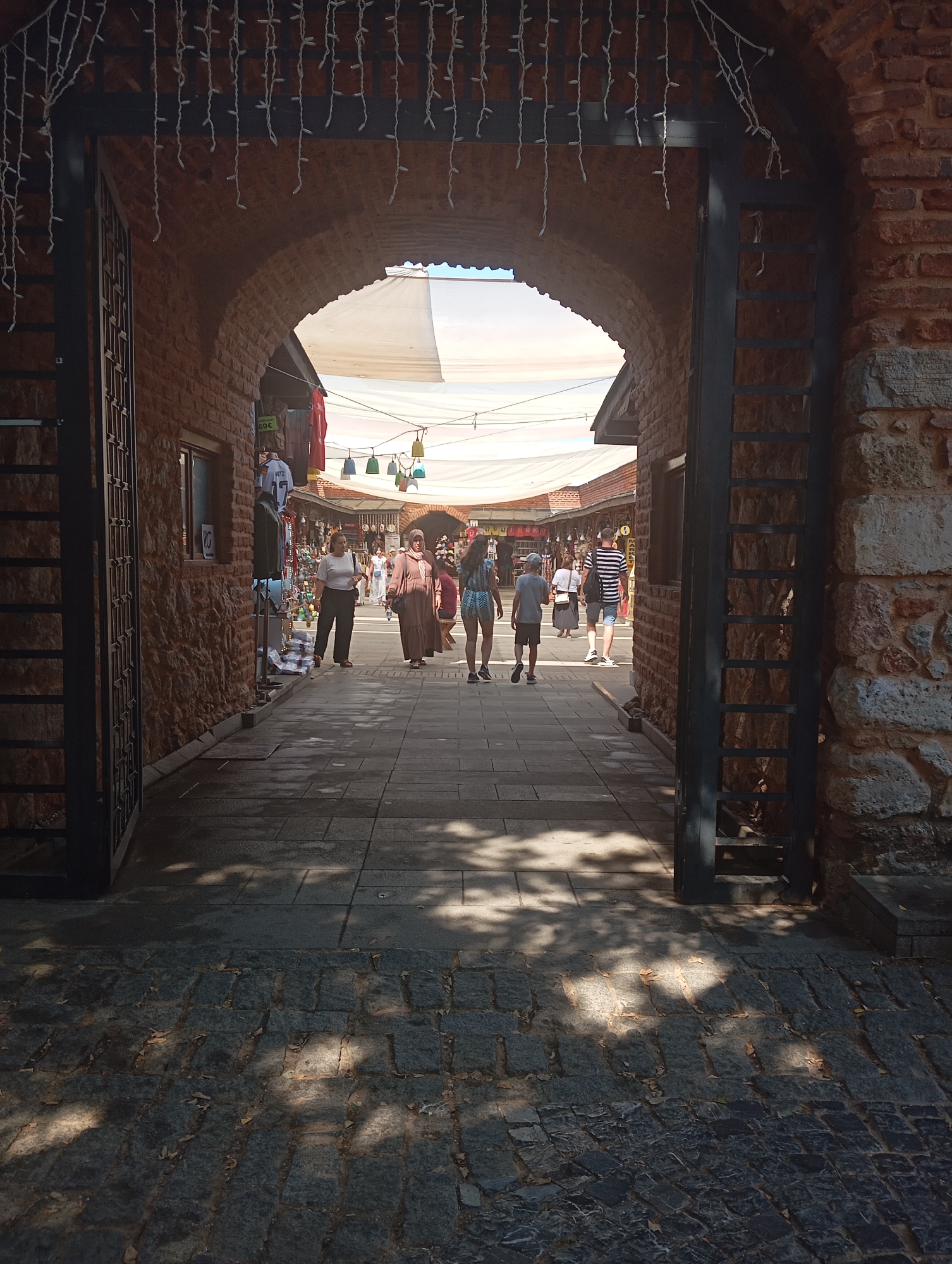 The image depicts an arched entryway leading into a marketplace in a Mediterranean-style city. The entryway is constructed of rough-hewn stone and brick, with a large metal gate on the left and right sides. Strings of white lights hang across the archway. Inside the marketplace, stalls are set up under white canopies, displaying various goods. Several people are visible browsing the stalls, including men, women, and children. The ground is paved with cobblestones. The lighting suggests it is daytime, with shadows cast by the sun. The overall impression is that of a bustling open-air market on a sunny day.