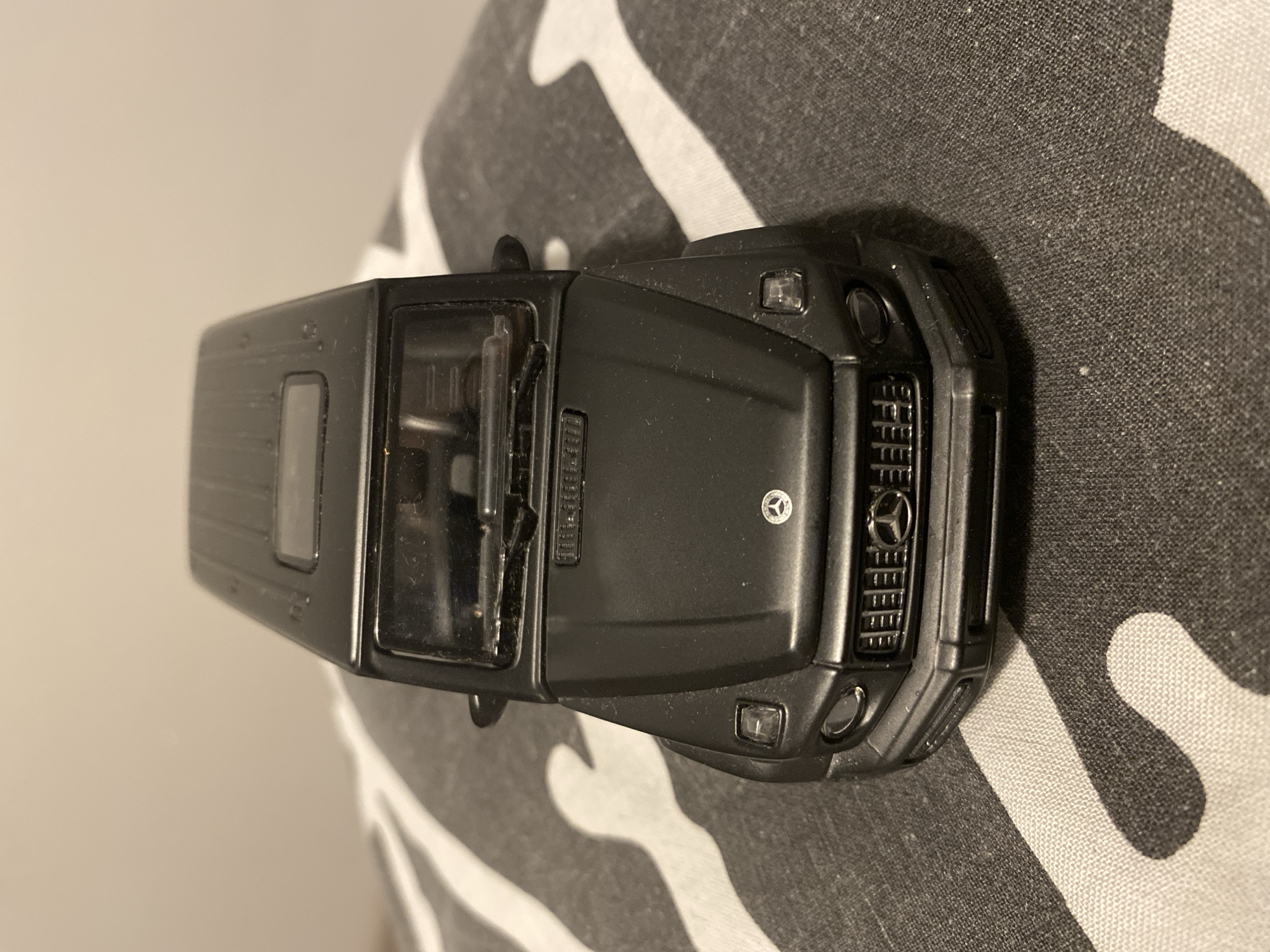 A black toy Mercedes-Benz G-Class is placed on a gray and white patterned fabric. The toy car is facing forward, with its headlights, grille, and hood clearly visible. The fabric appears to be a textile with a repeating abstract pattern. The background is a plain, light-colored wall. The scene is likely indoors, on a flat surface. There are no people or animals present in the image. The image does not contain any information that would allow for the determination of a location in the city of Łódź, Poland.
