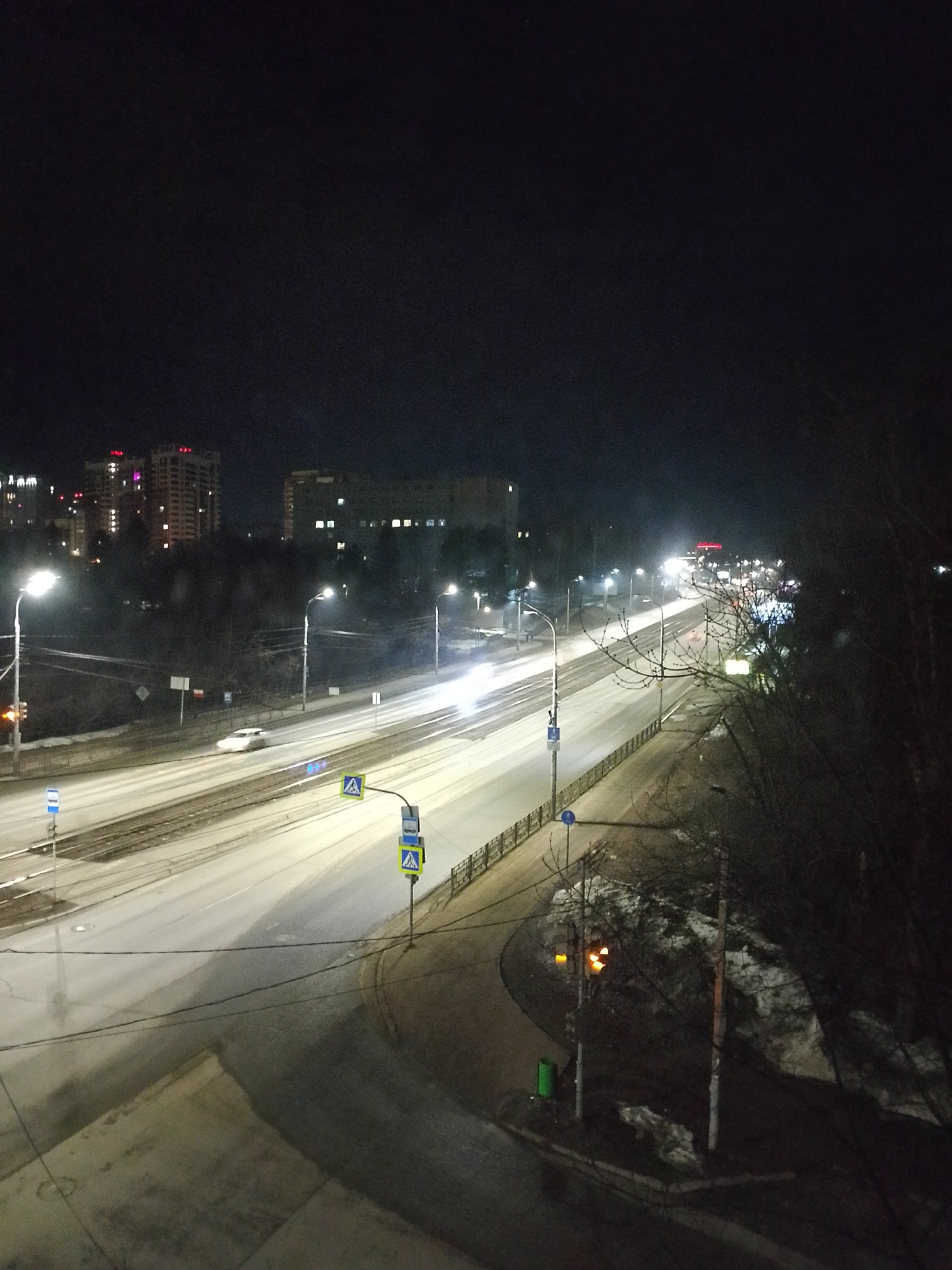 The image depicts a nighttime urban scene in Izhevsk, Russia. A wide, multi-lane road dominates the foreground, illuminated by streetlights and vehicle headlights. The road appears wet or icy. Sidewalks and pedestrian walkways are visible on either side of the road. In the background, tall buildings with illuminated windows suggest a residential or commercial area. Trees line the roadway. Traffic signs and road markings are present. A small patch of snow is visible near the roadside. A green trash can is also visible. Bare trees stand to the right.